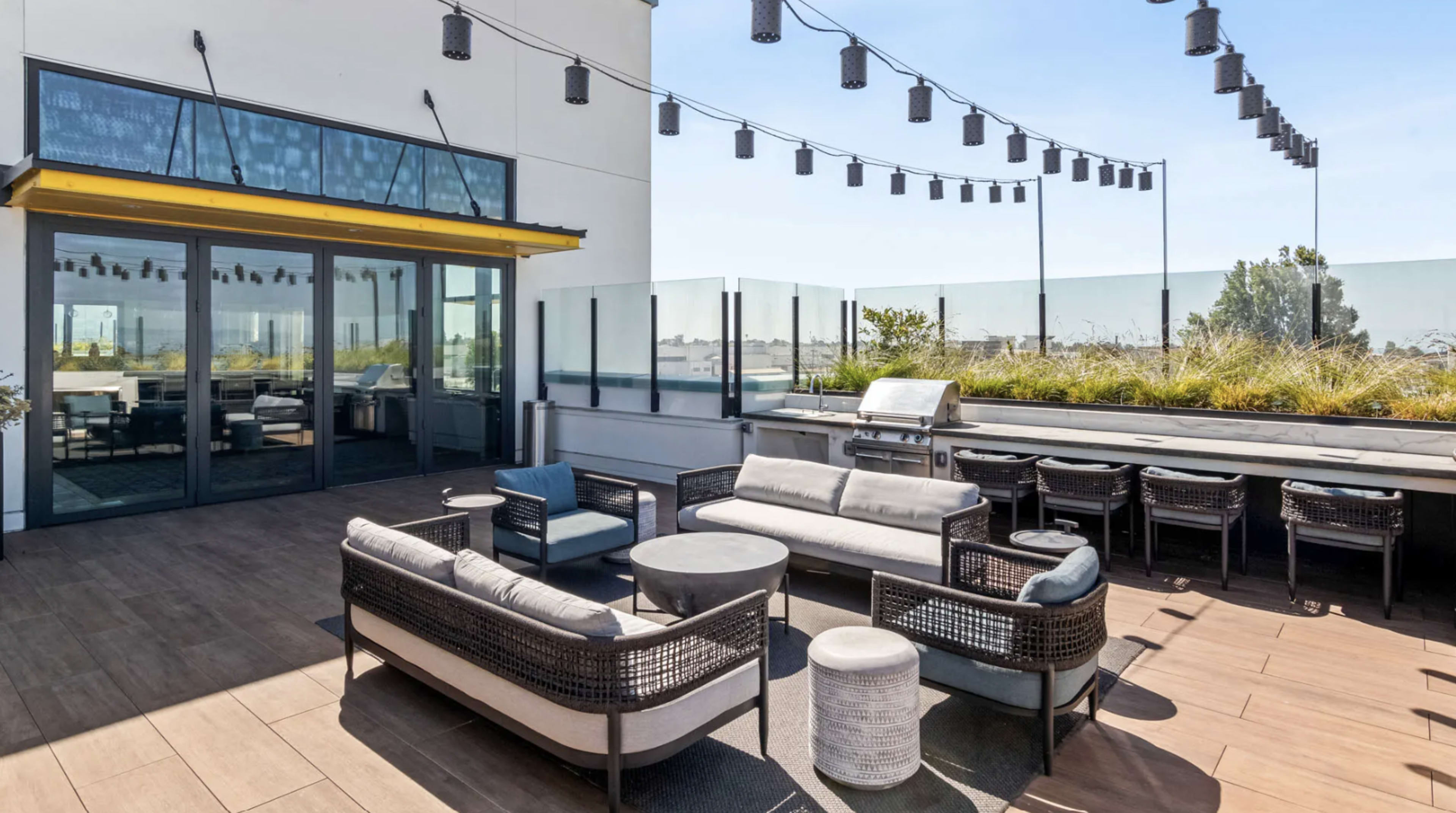 The image shows a modern outdoor patio area with comfortable seating, a dining space, and string lights overhead.