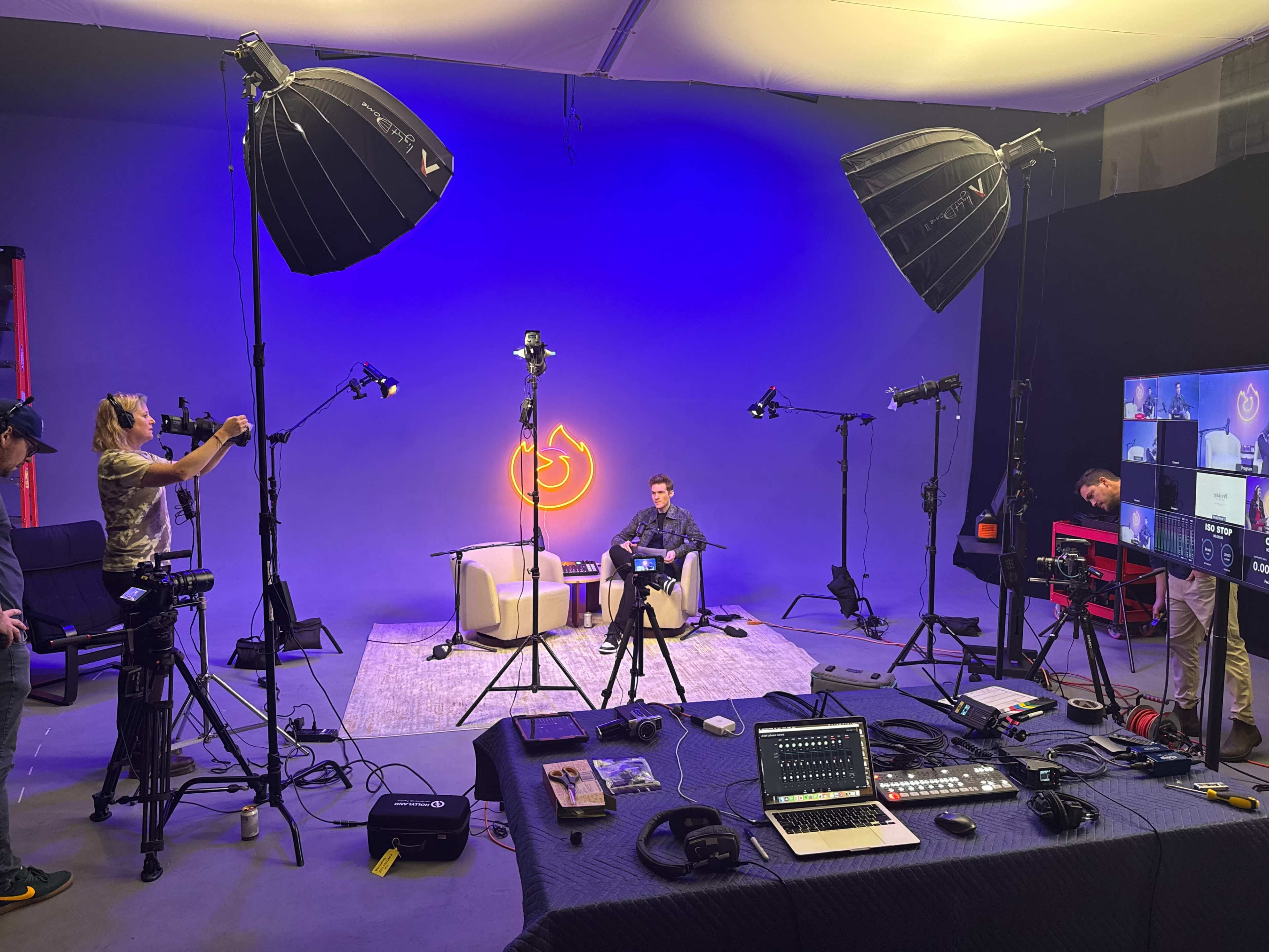 The image shows a studio setup with multiple cameras and lighting equipment surrounding a seating area where a person is sitting, and screens displaying a production interface are visible in the background.