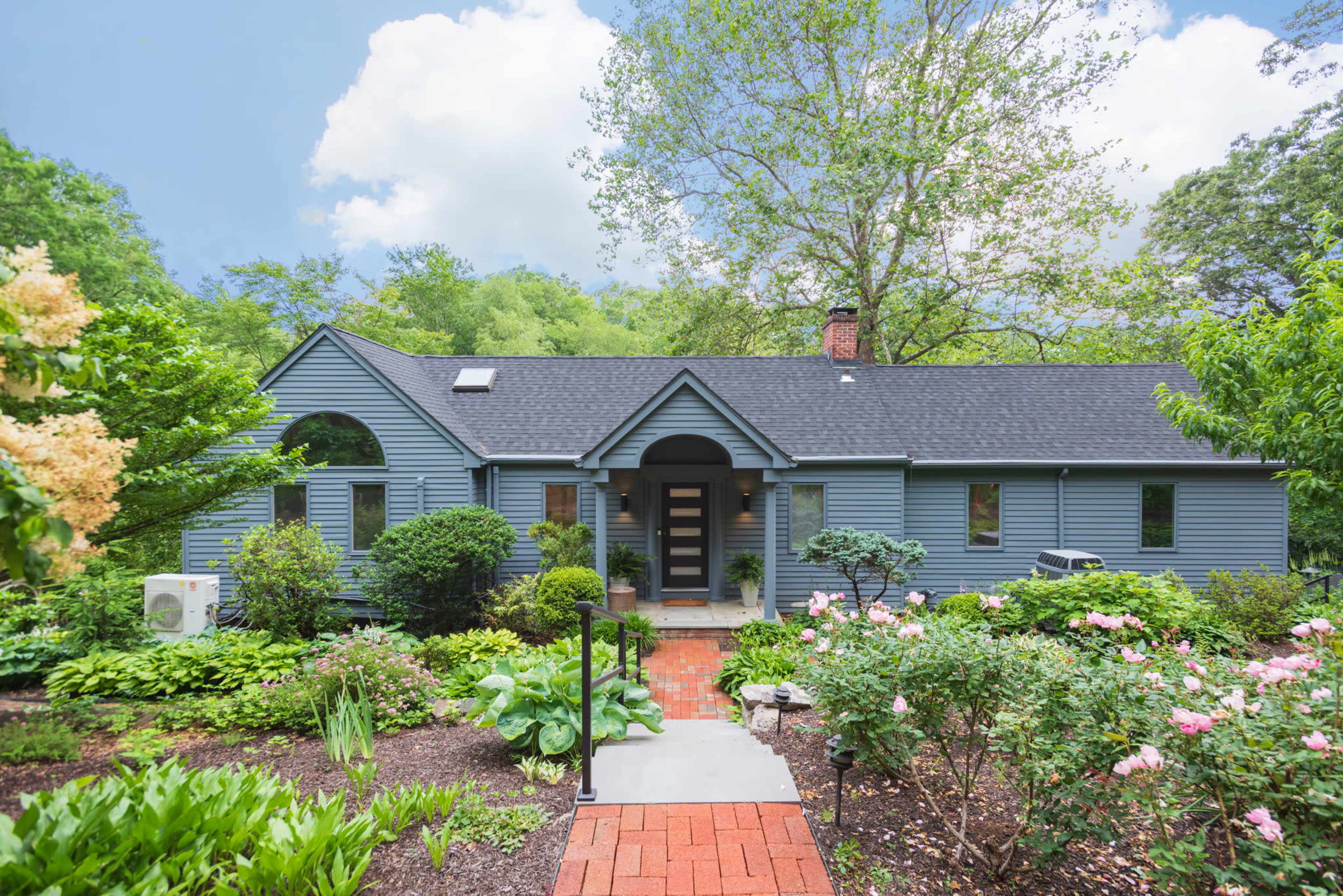 A blue residential house is set amidst lush greenery and well-maintained flowerbeds, with a brick pathway leading to the entrance.