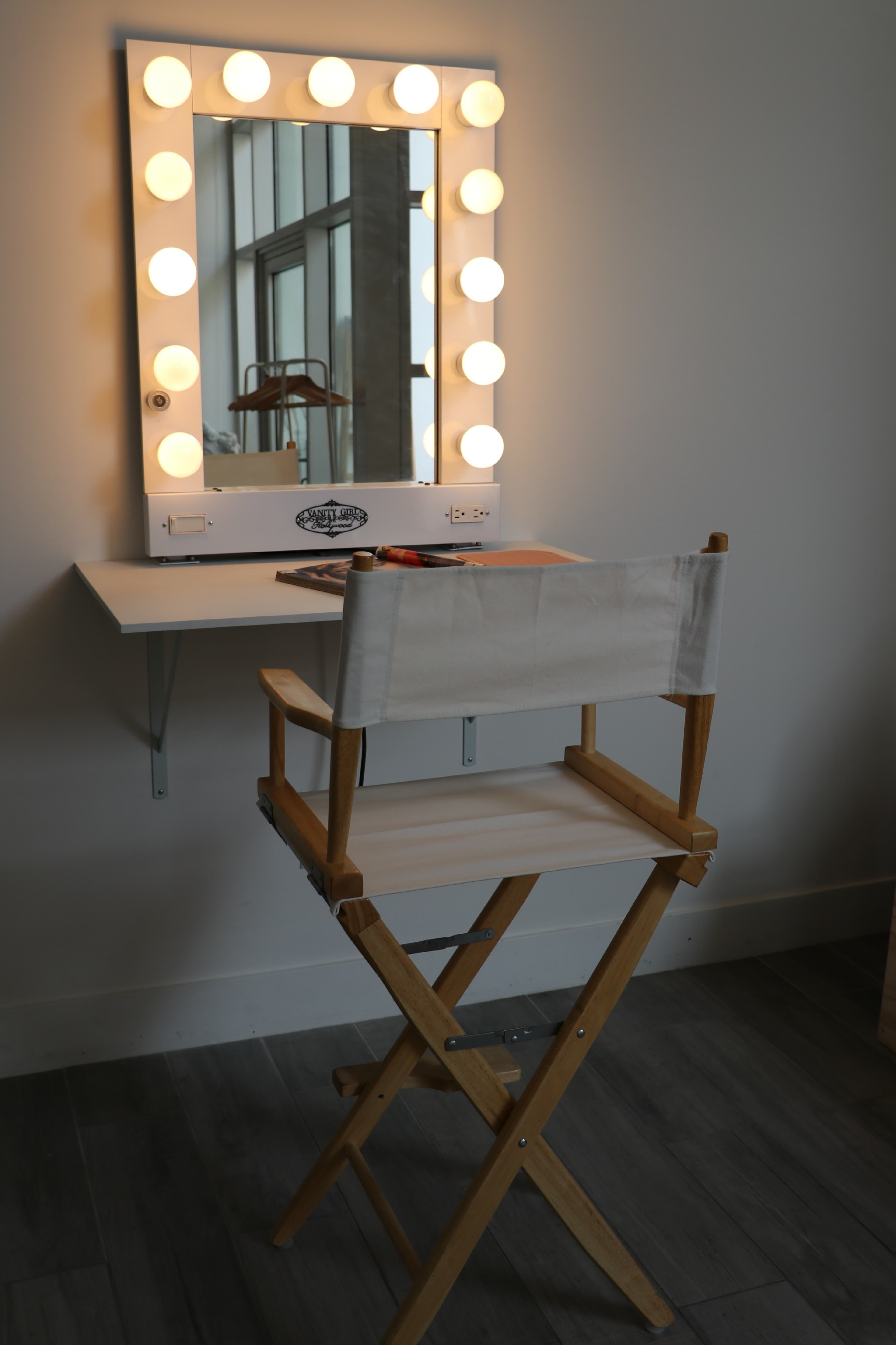 A brightly lit makeup mirror is mounted on the wall above a wooden director's chair in a minimalist room.