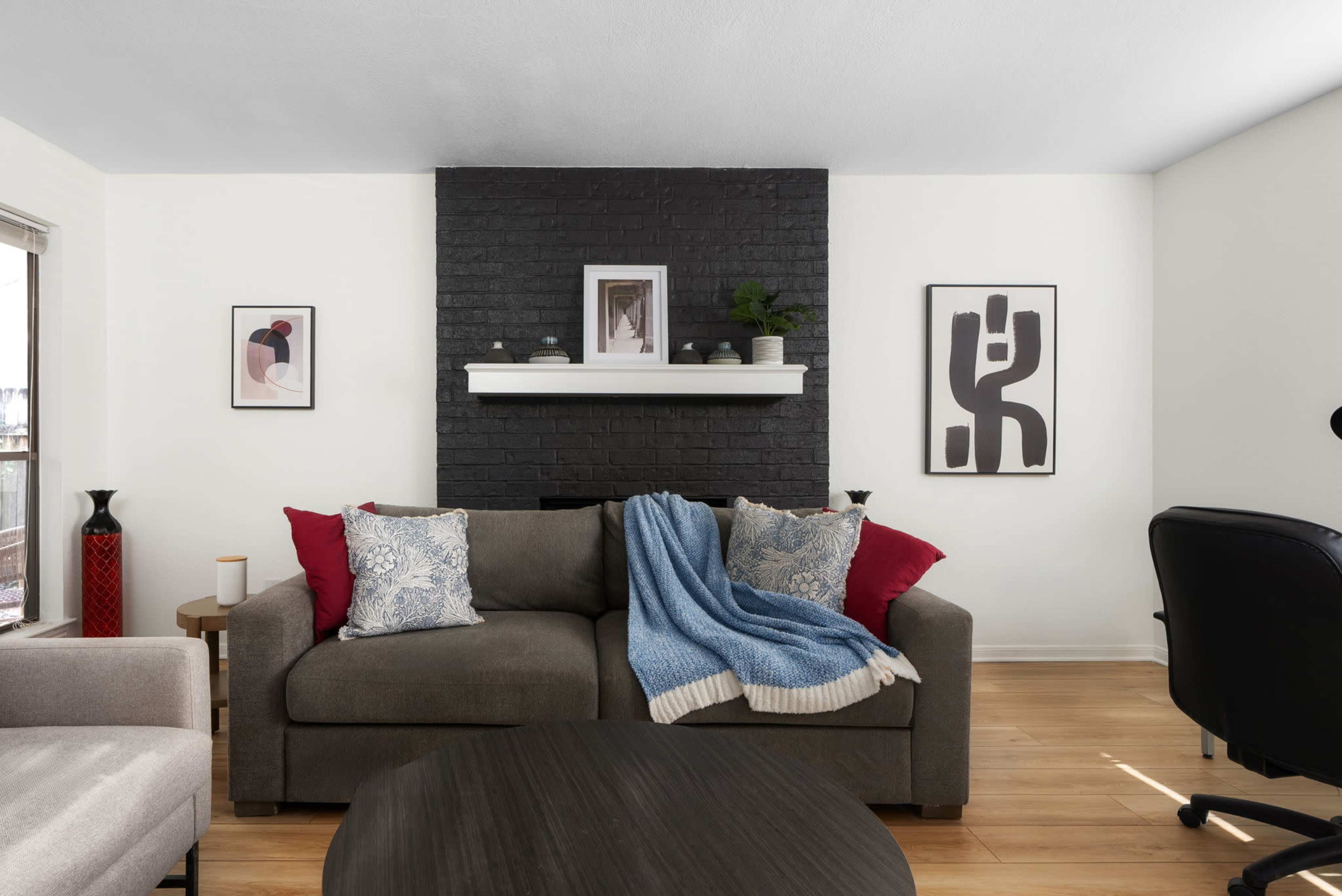 A cozy living room features a dark brick fireplace with decorative artwork above it, a gray sofa adorned with colorful pillows and a blue throw blanket, and a round coffee table.