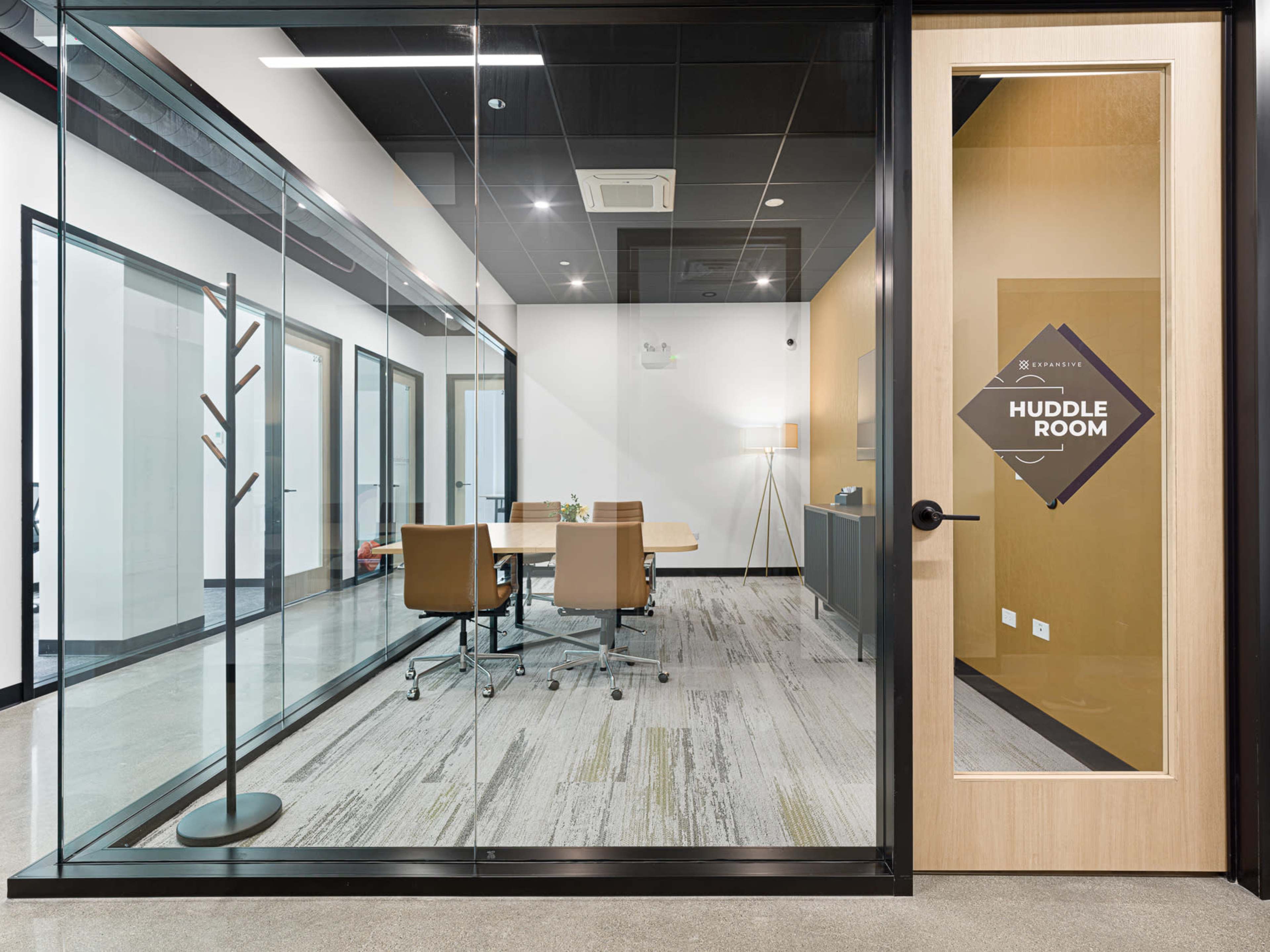 The image shows a modern huddle room with a glass wall, a wooden door, and a table surrounded by four chairs inside.