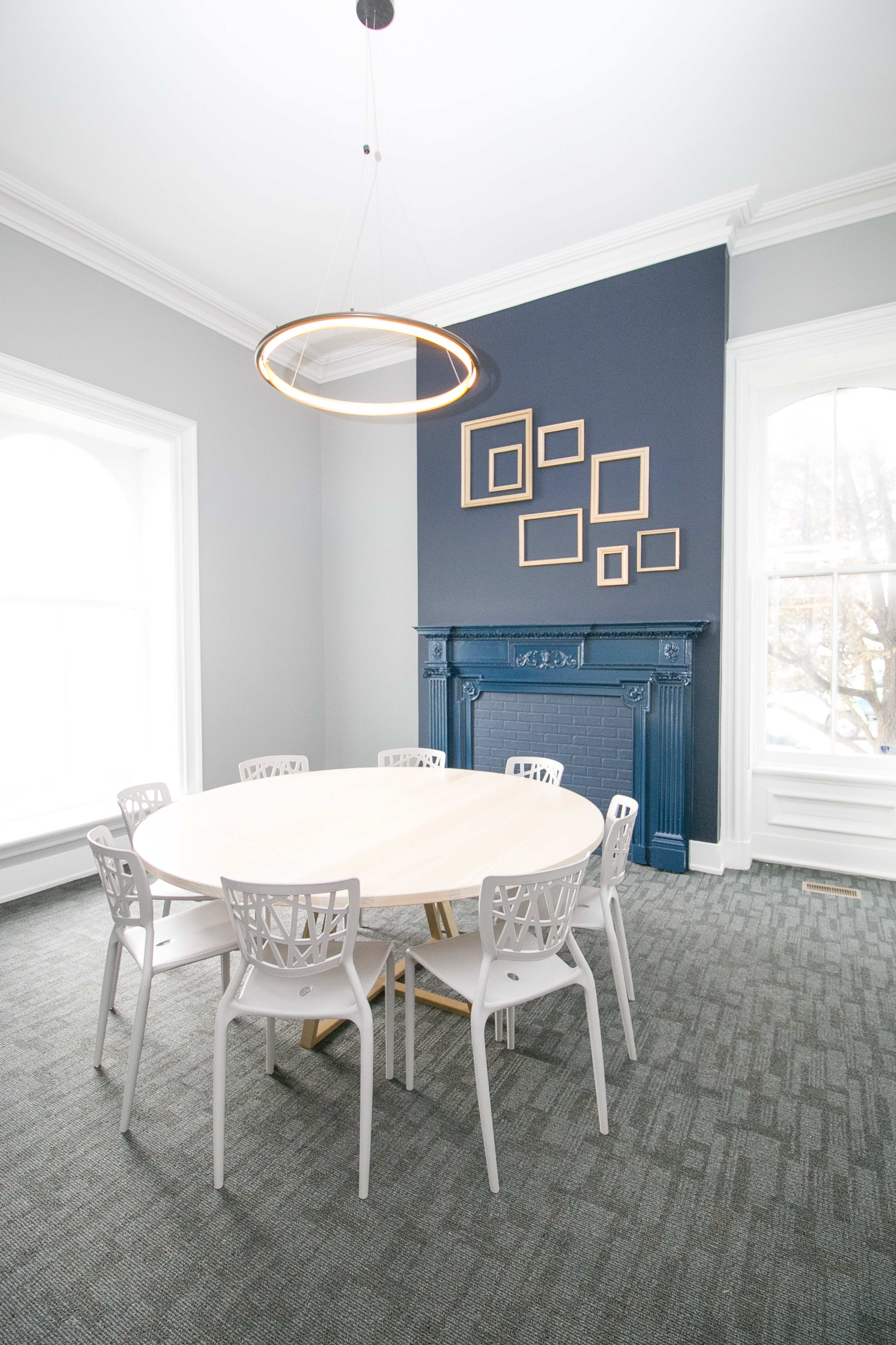A modern meeting room with a circular wooden table surrounded by white chairs and a dark blue accent wall featuring an ornate fireplace and several empty picture frames.