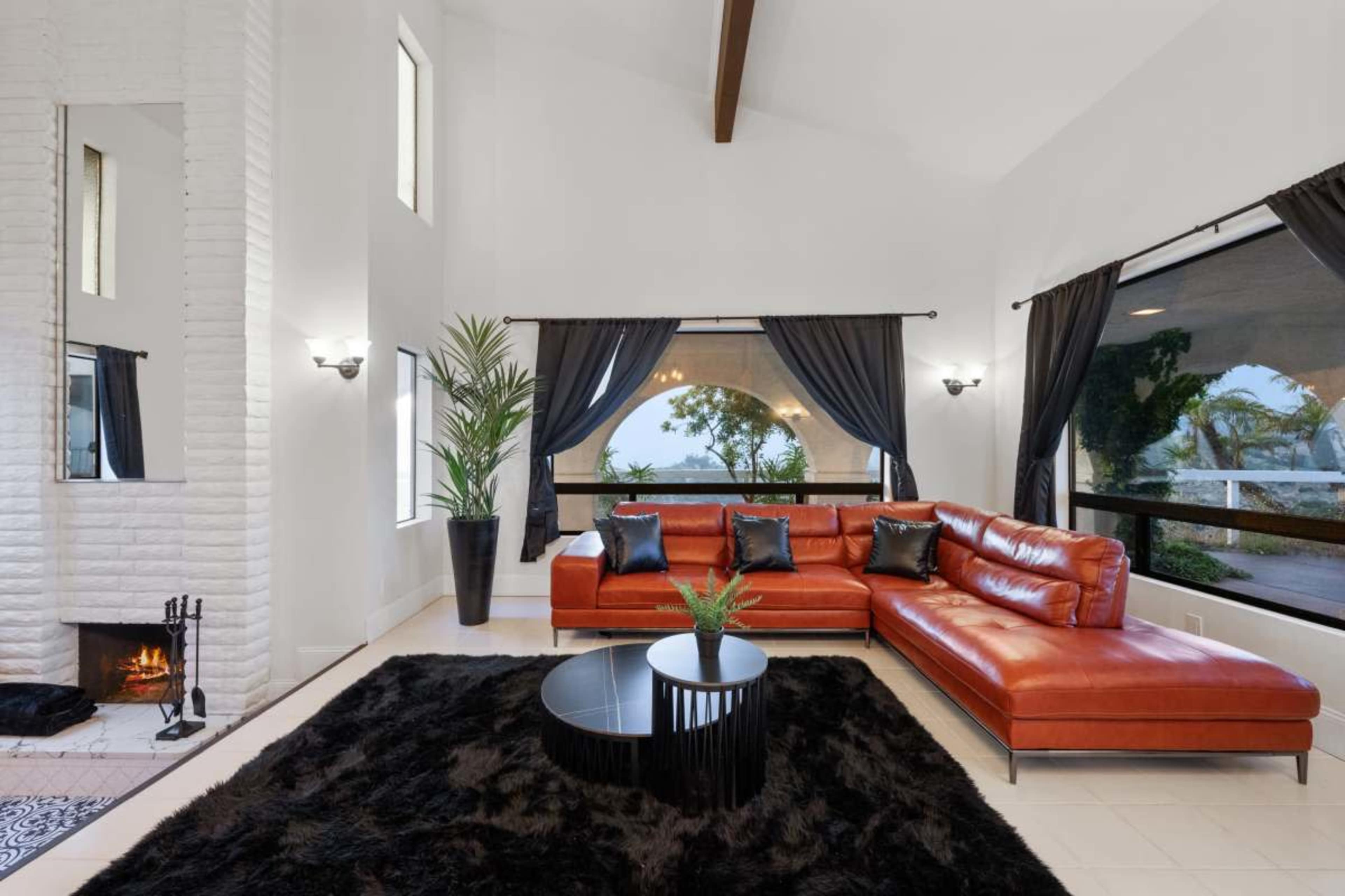 The image shows a modern living room featuring a large red leather sectional sofa, a circular black coffee table, and a black rug, with large windows providing a view of the outdoors.