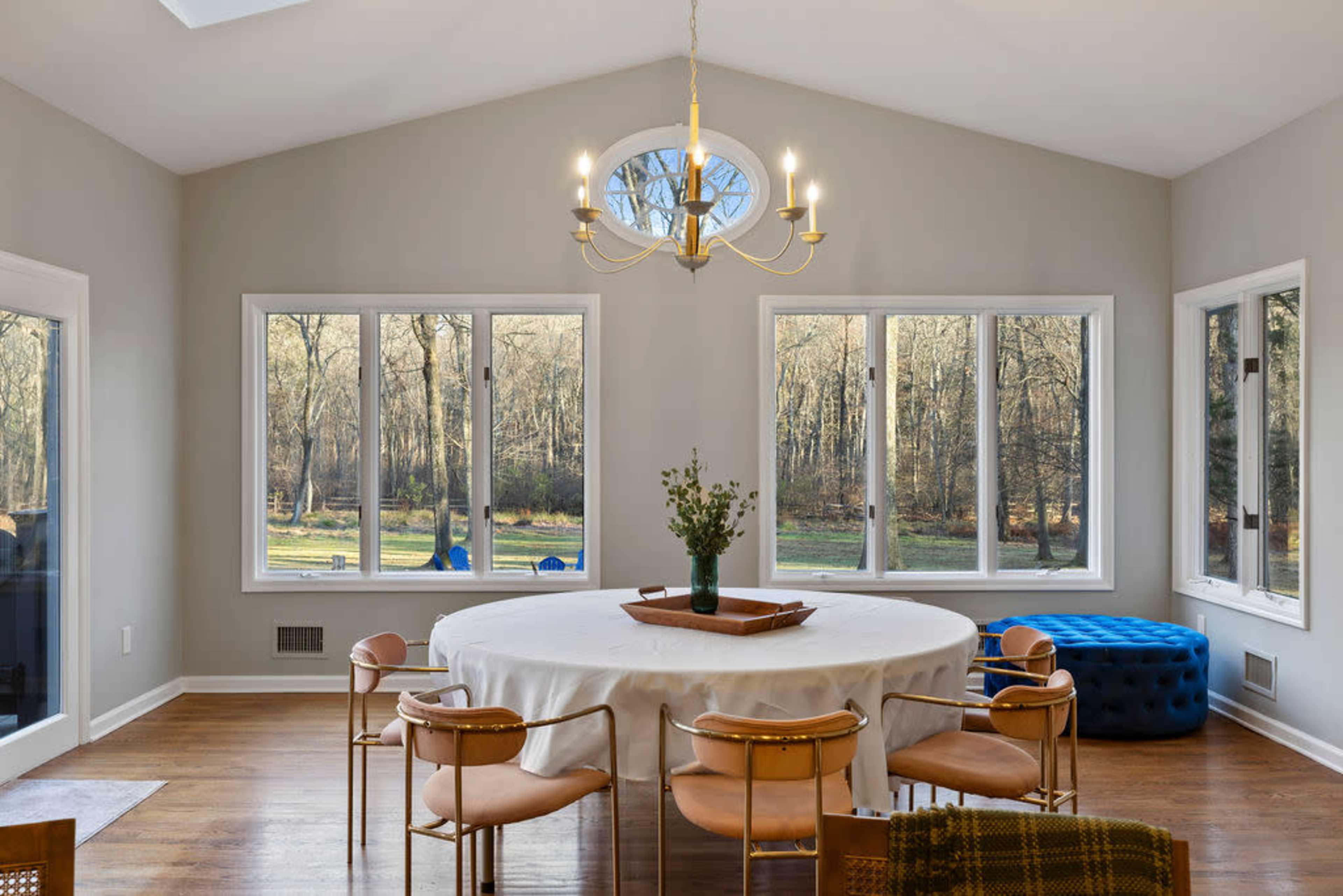 A circular dining table surrounded by eight chairs is situated in a brightly lit room with large windows overlooking a forested area.
