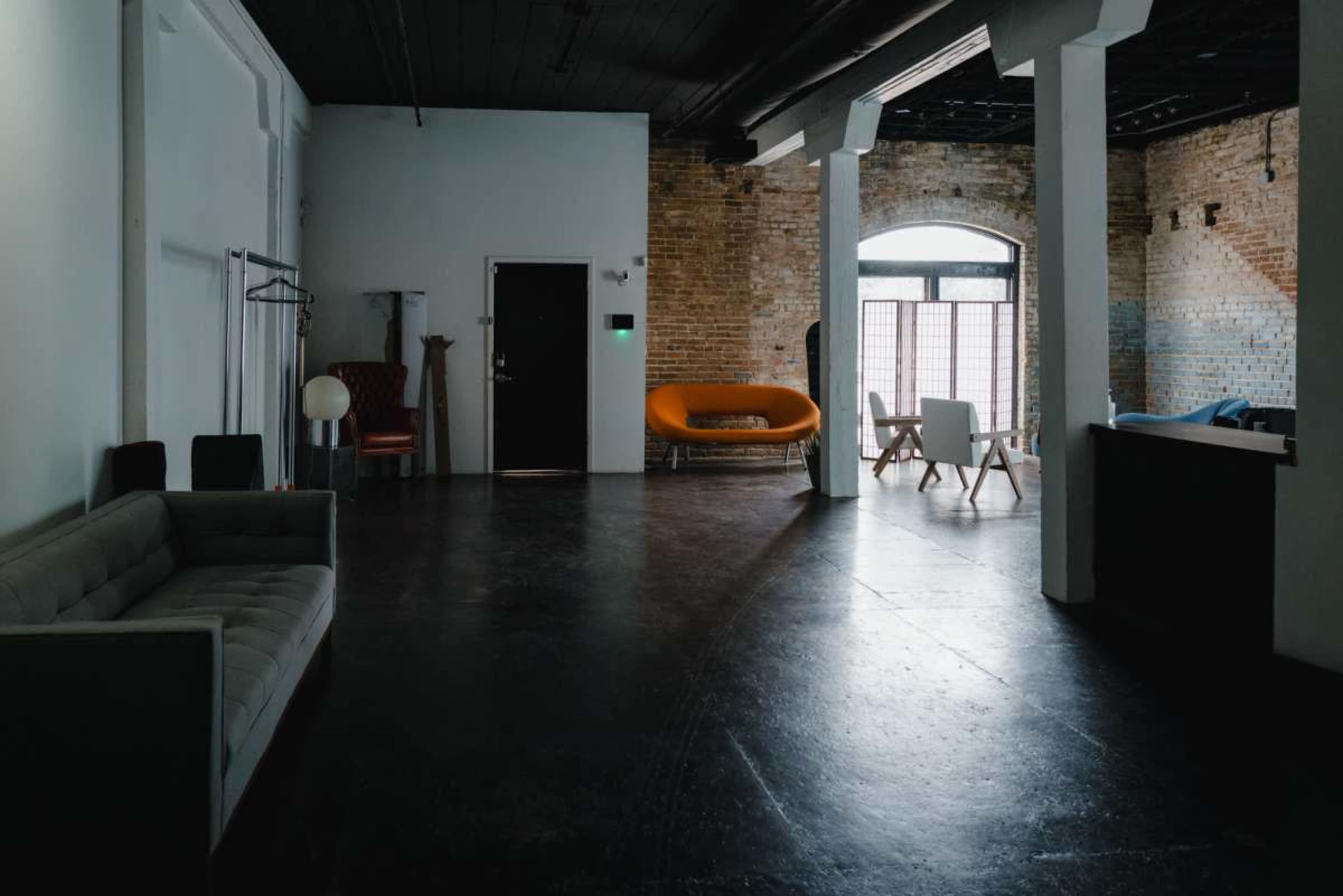 The image shows a spacious, minimalist room with exposed brick walls, featuring a gray sofa, an orange couch, and various furniture arranged in different areas.