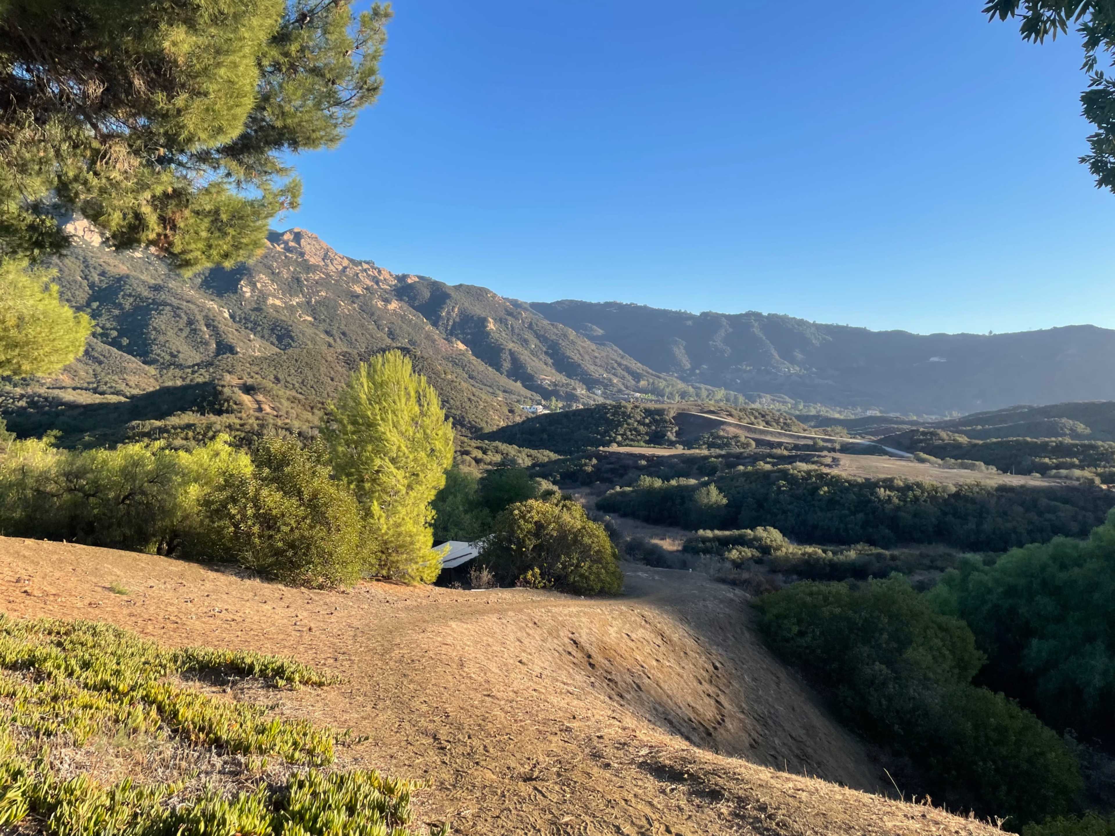 The image shows a mountainous landscape with rolling hills and a clear blue sky, dotted with trees in the foreground.