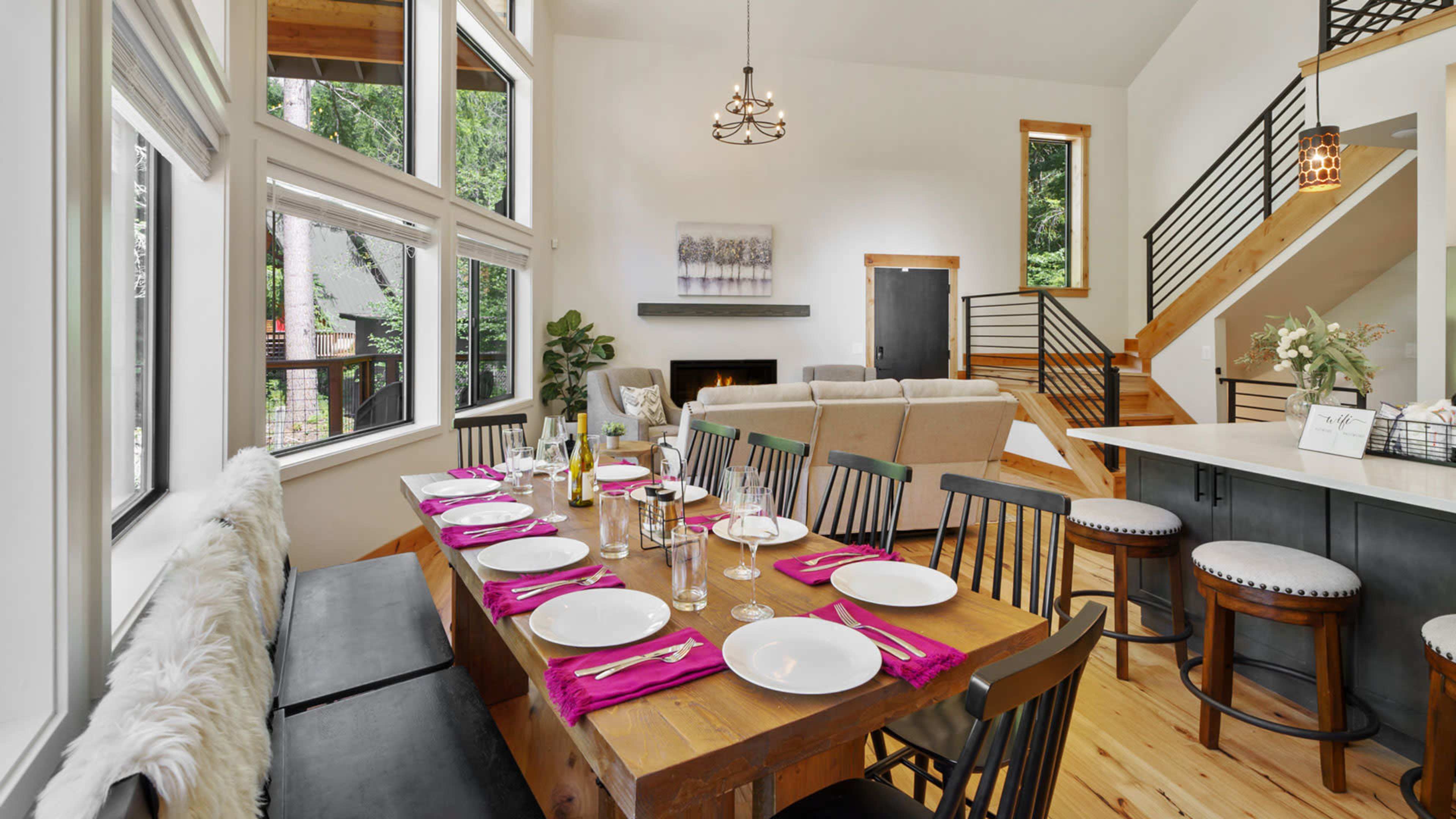 A dining area with a wooden table set for a meal, surrounded by large windows and a staircase leading to an upper level.