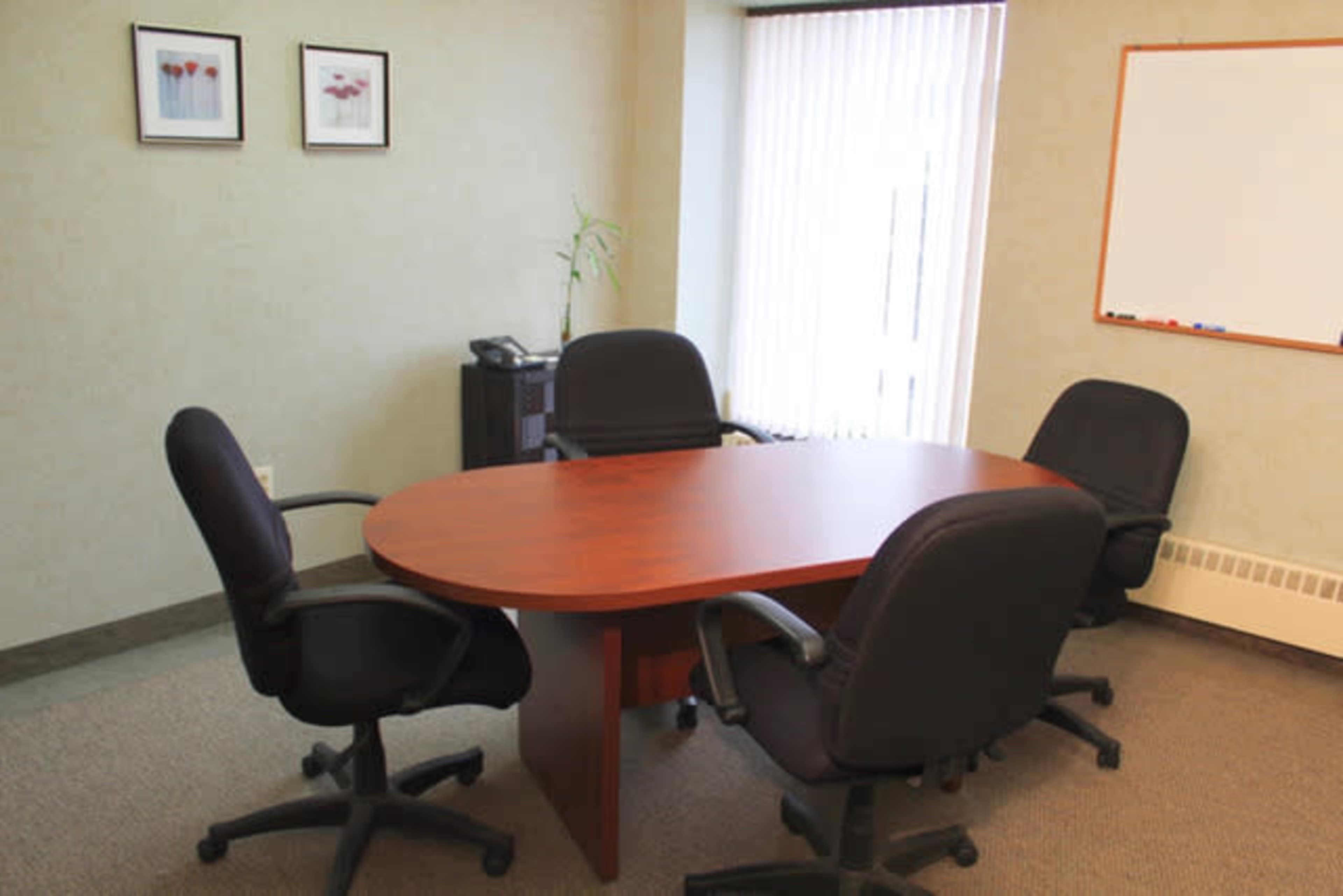 A conference room features a wooden oval table surrounded by four black office chairs, with a whiteboard and two framed pictures on the wall.