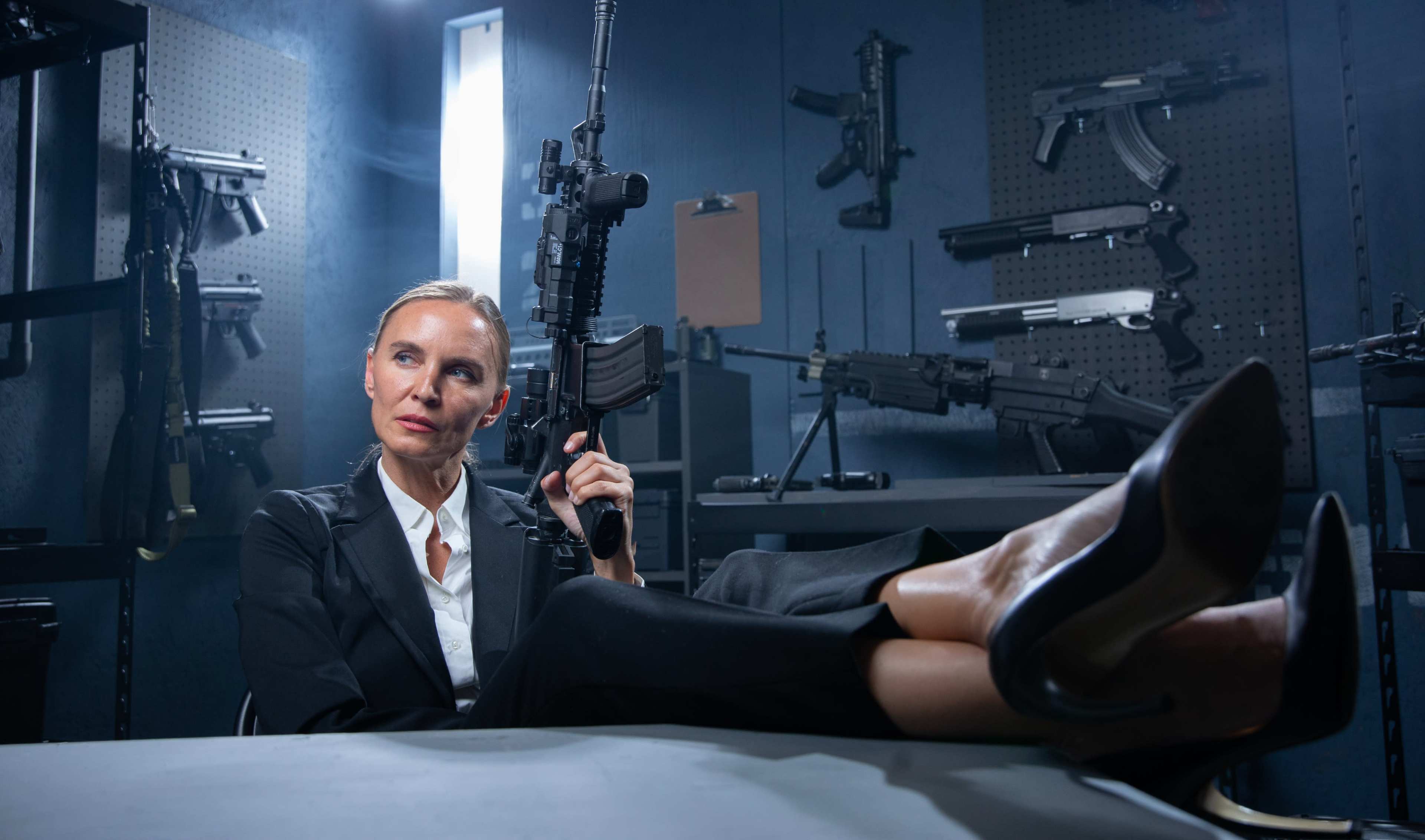 A woman in a suit poses with an assault rifle while reclining at a table in a dimly lit room filled with various firearms.
