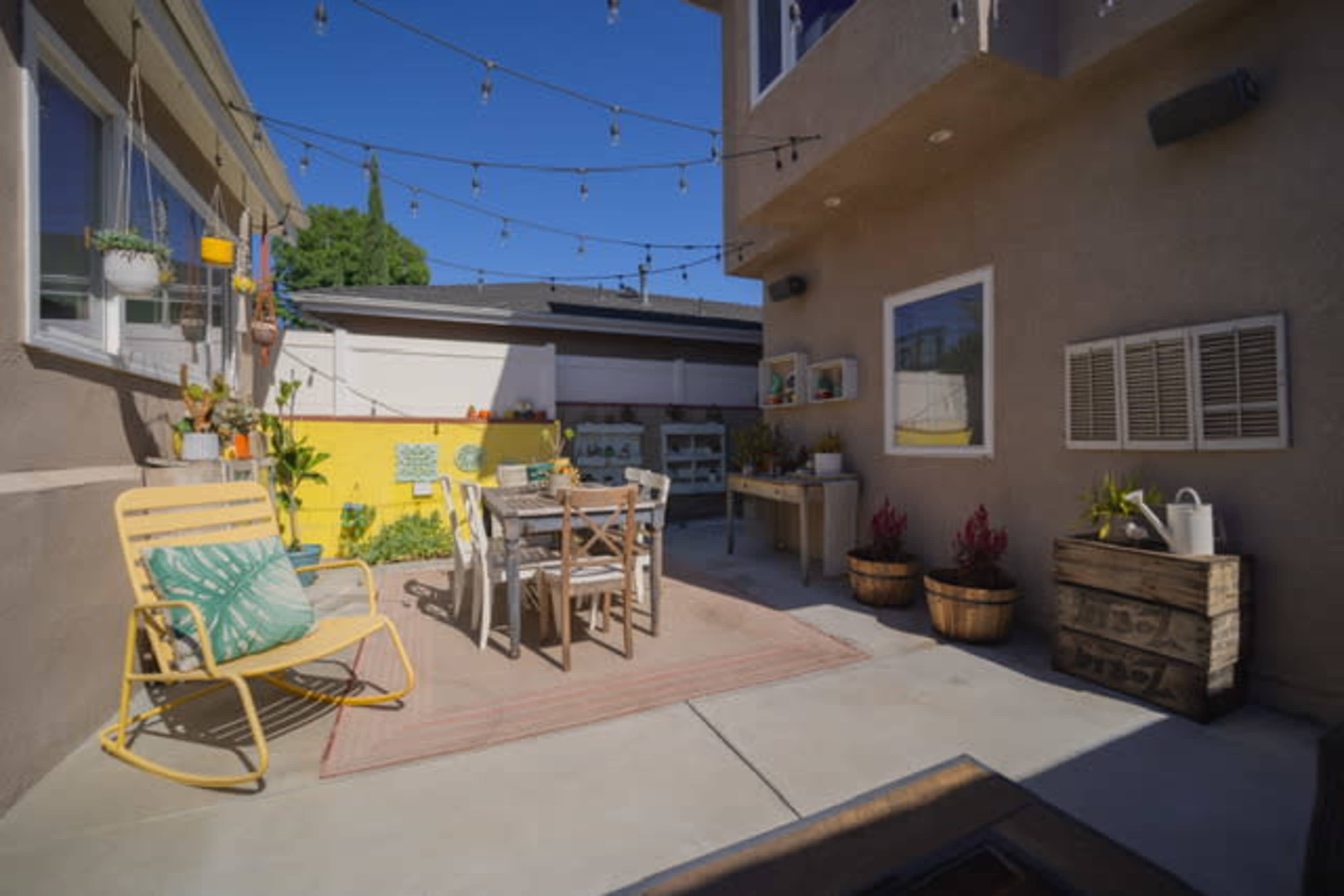 A patio area with a dining table, chairs, and string lights, bordered by yellow walls and various plants.