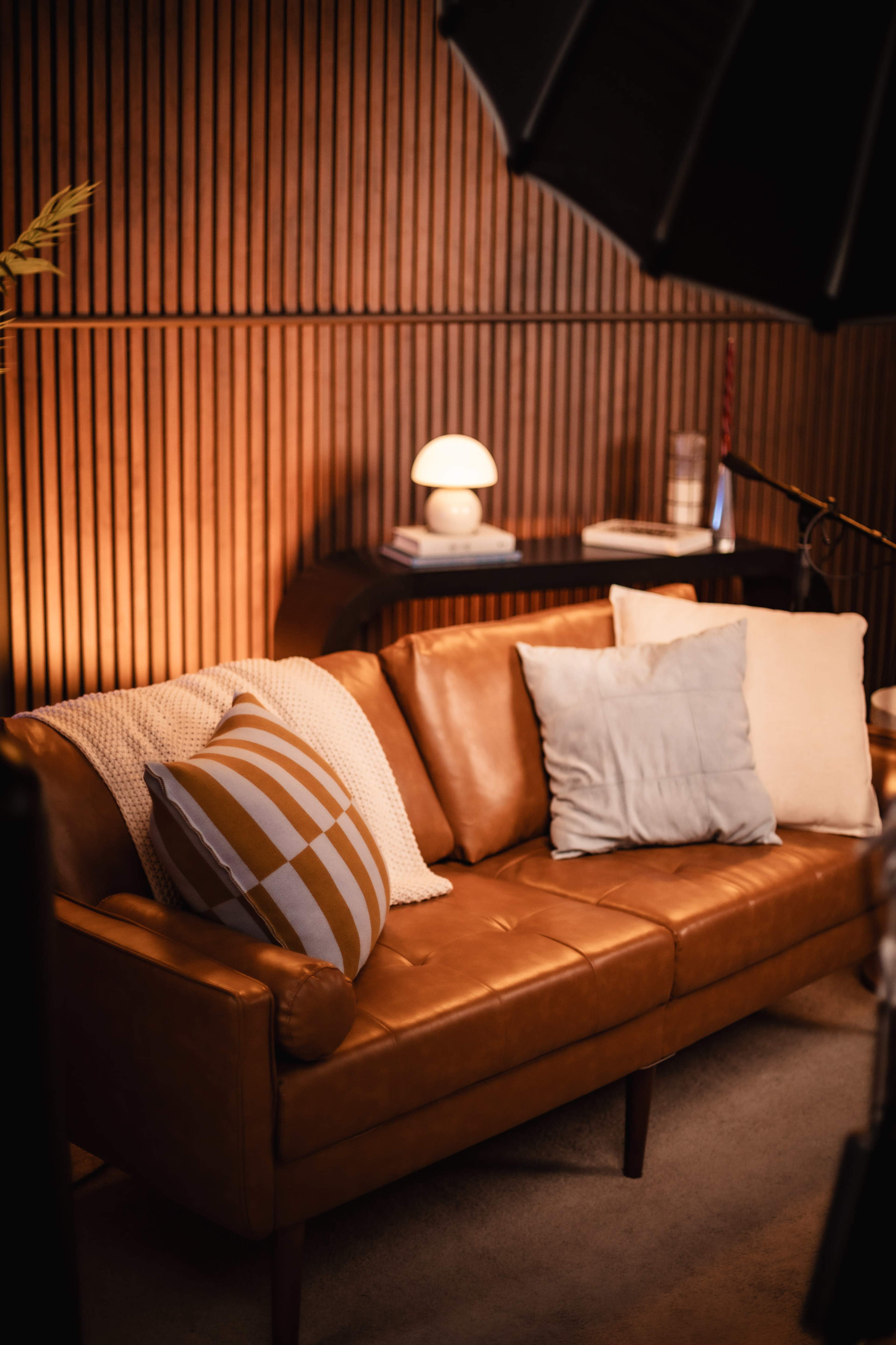 A brown leather sofa with various cushions is positioned in a warmly lit room featuring wooden paneling.