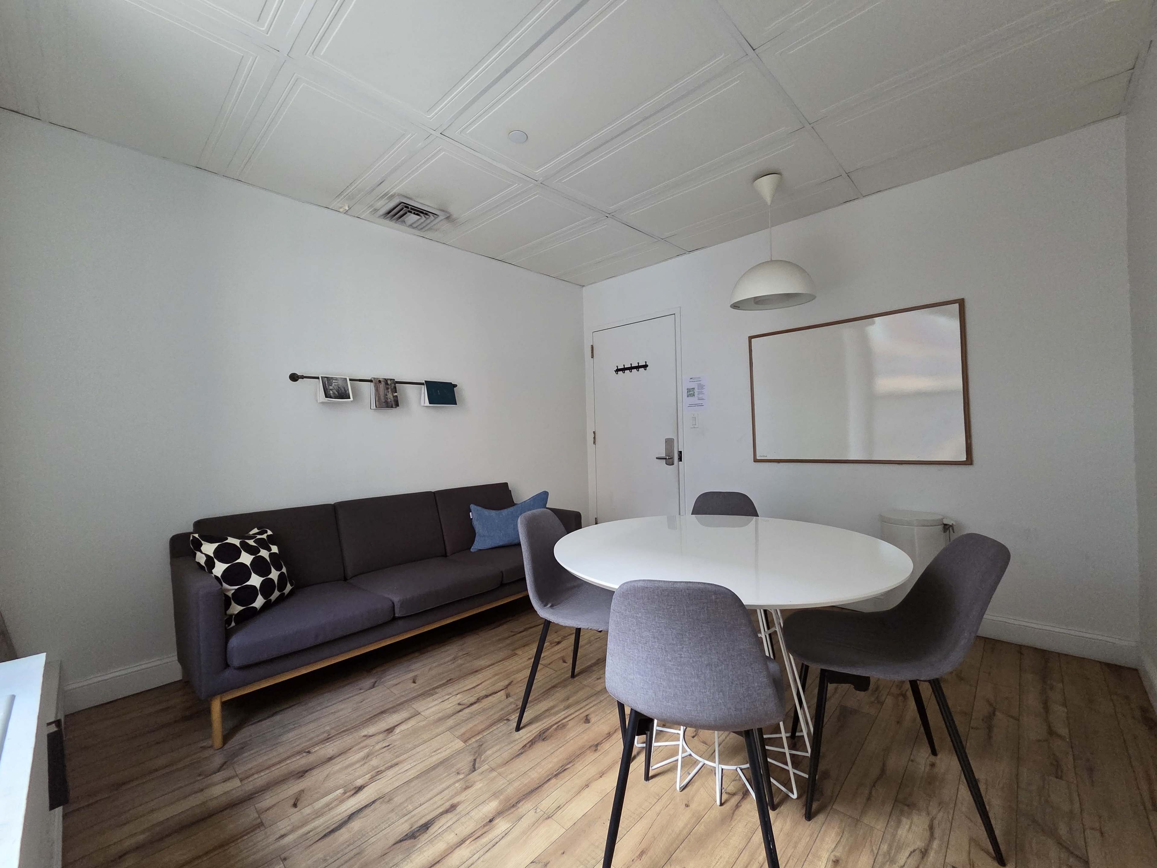 The image shows a small room featuring a gray sofa with decorative pillows, a round white dining table surrounded by gray chairs, and a light fixture hanging from the ceiling.