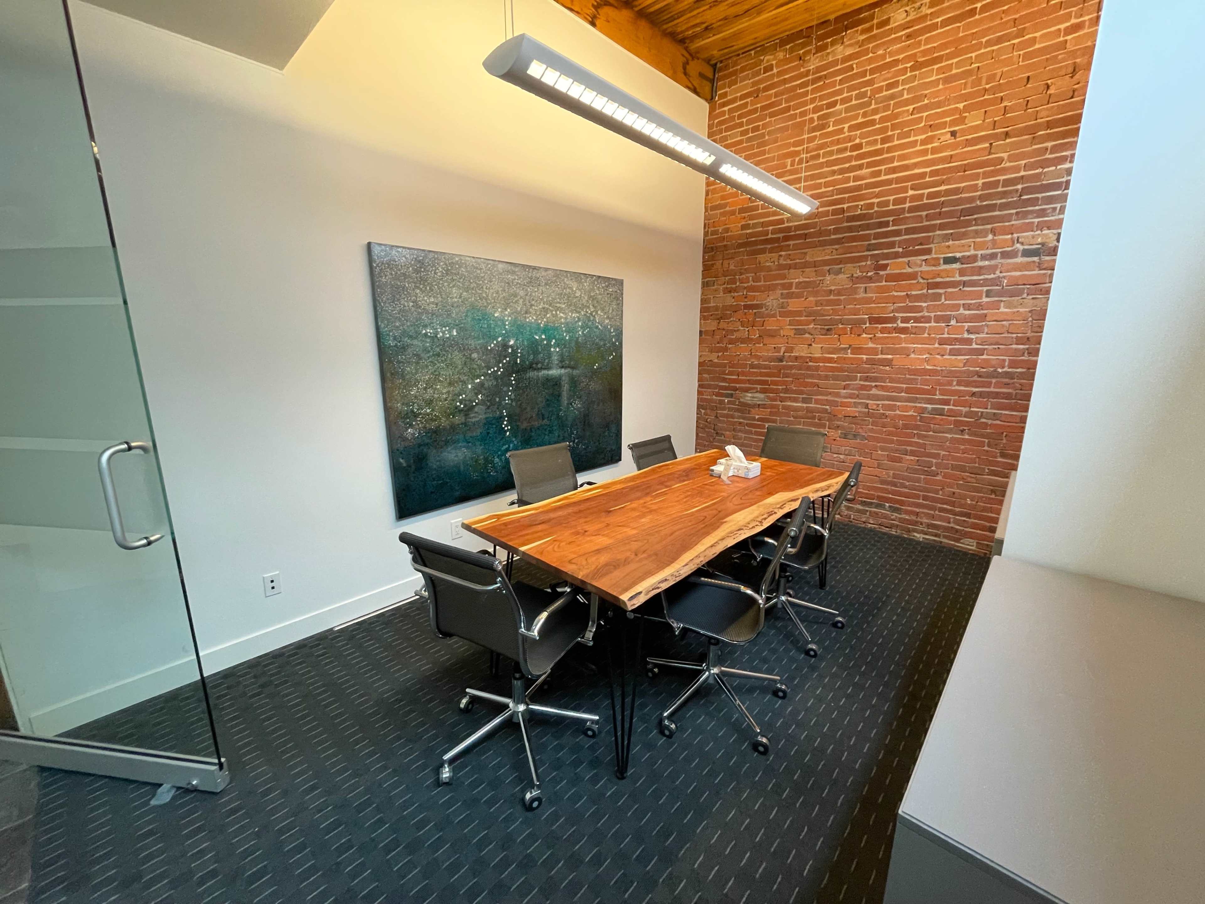 A conference room features a wooden table surrounded by chairs and a large abstract painting on the brick wall.