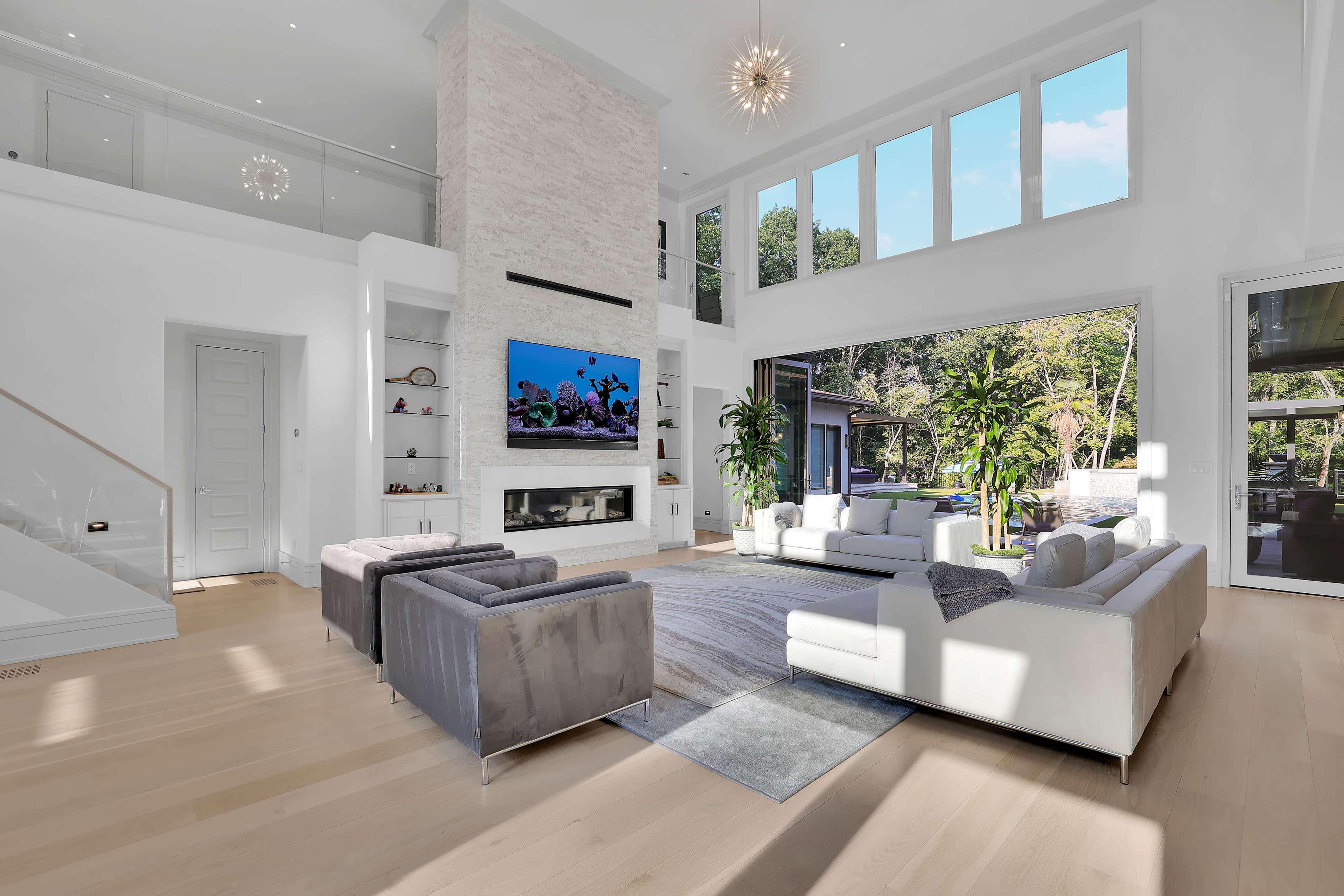 A bright, modern living room features large windows, minimalist furniture, and a fireplace with a TV mounted above it.