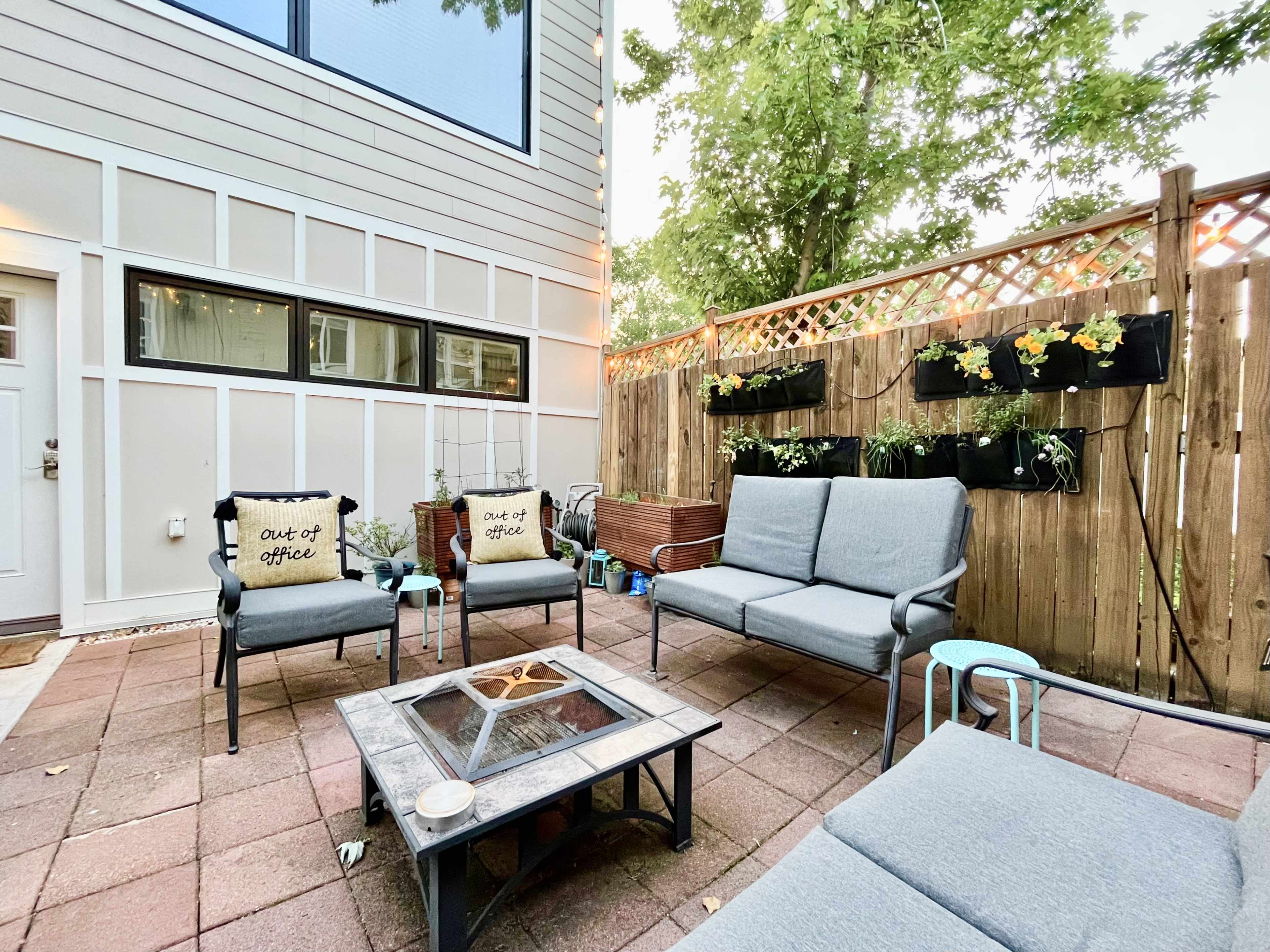 A patio area features a seating arrangement with gray cushions, a fire pit in the center, and potted plants along the fence.