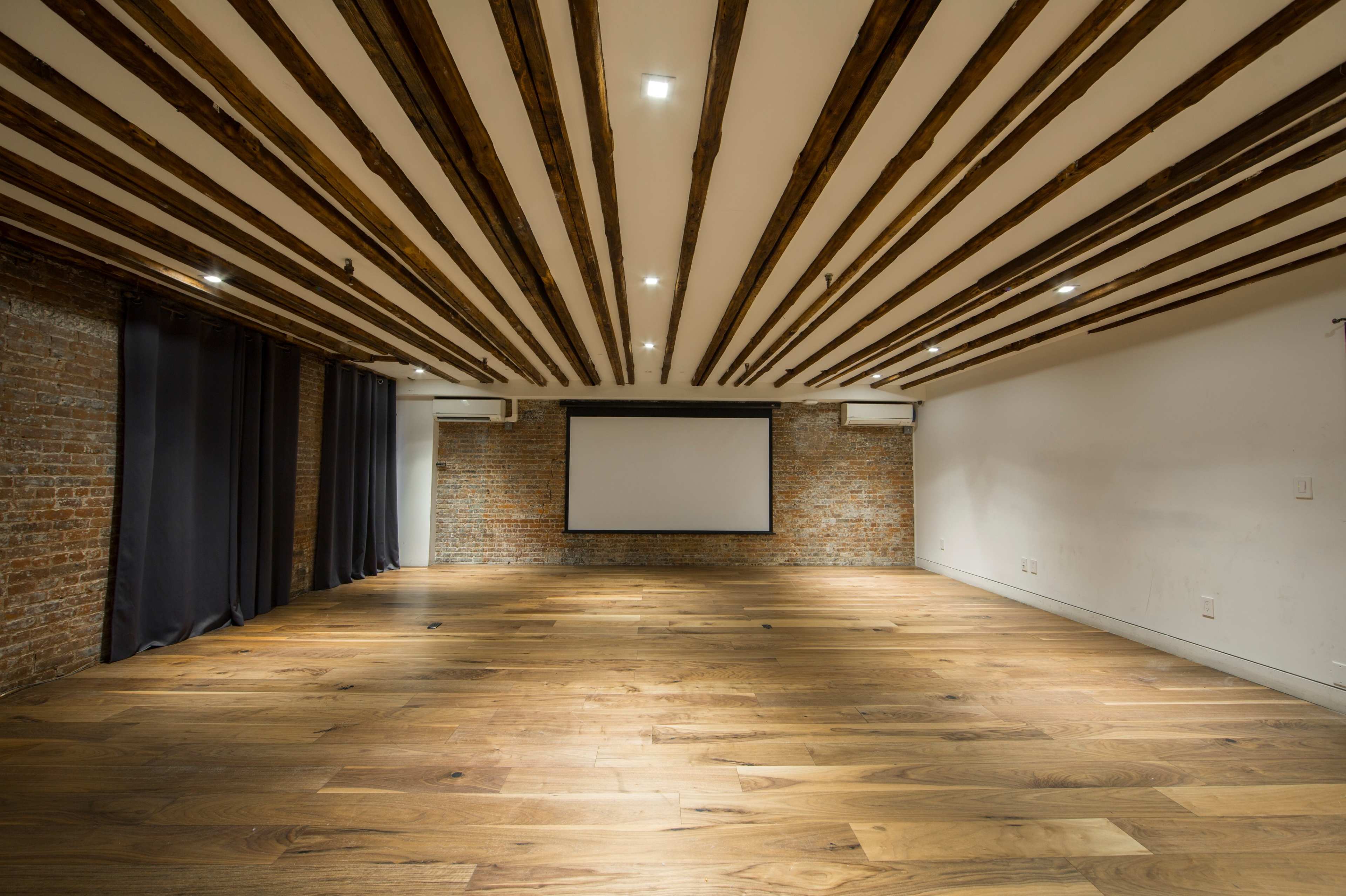 A spacious room with wooden beams on the ceiling, brick walls, a blank projector screen, and wide wooden flooring.