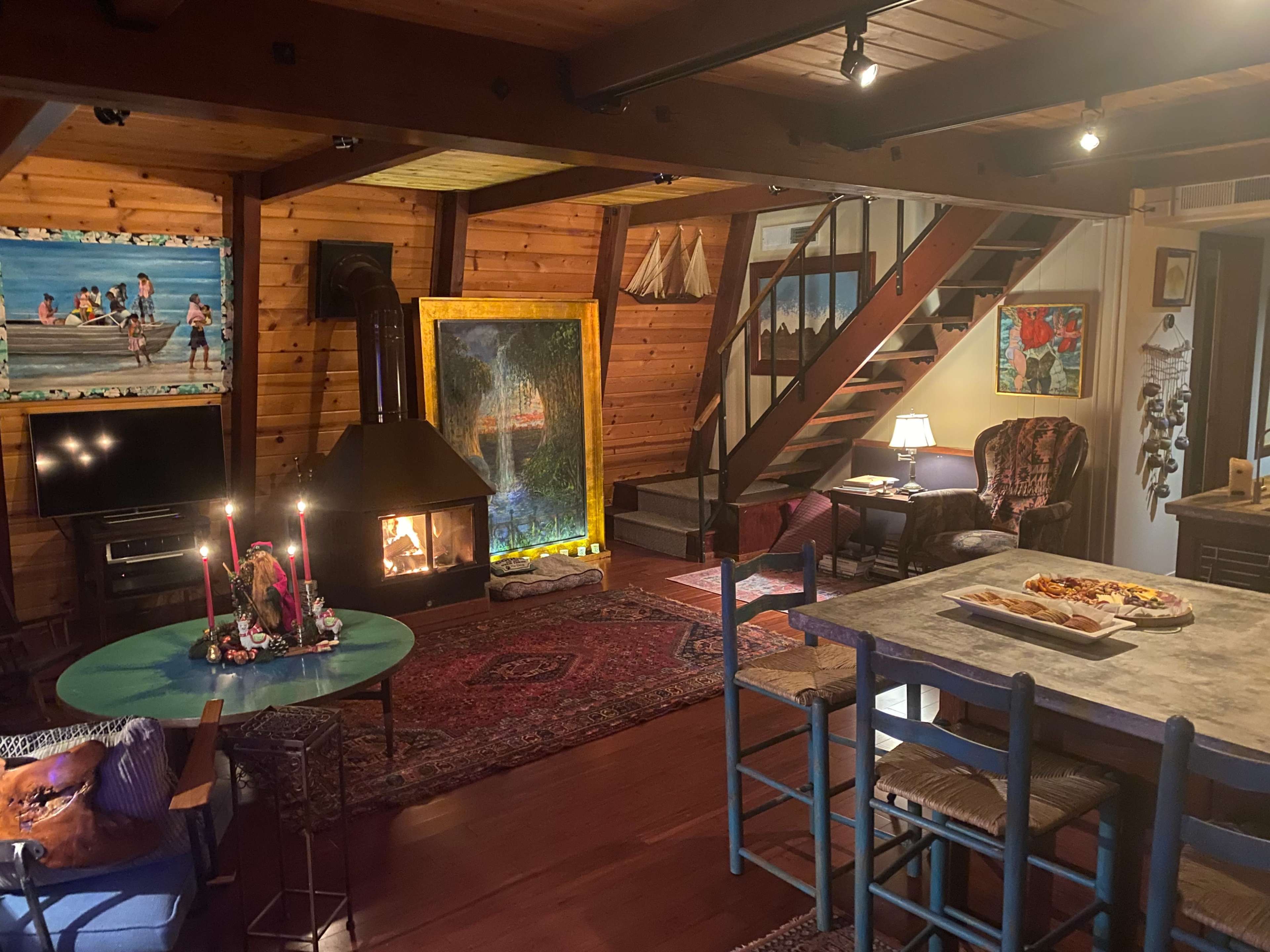 A cozy interior of a wooden cabin featuring a fireplace, a dining table, stairs, various artworks, and comfortable seating arrangements.