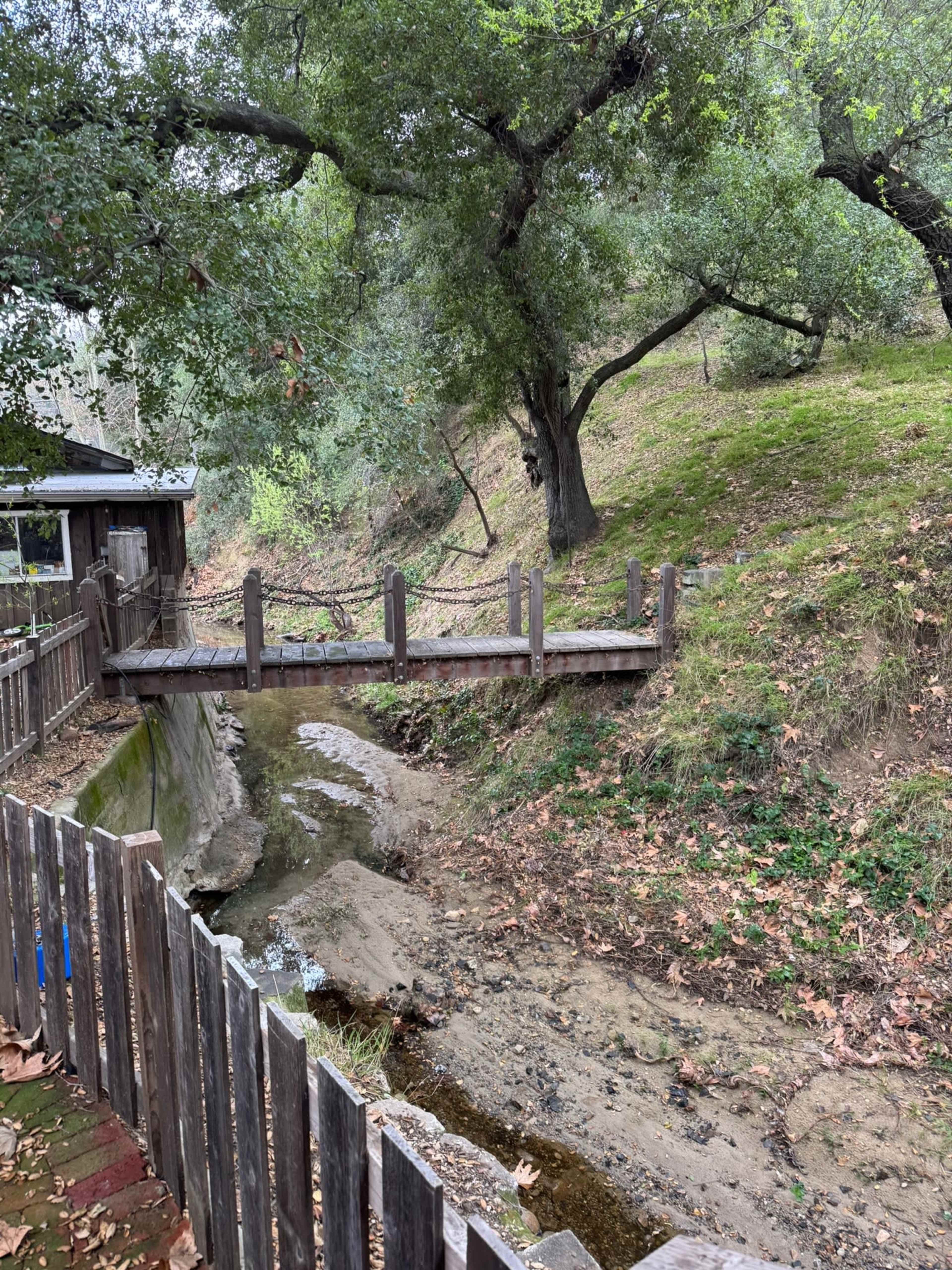 A wooden bridge crosses a dry creek bed surrounded by grassy slopes and trees.