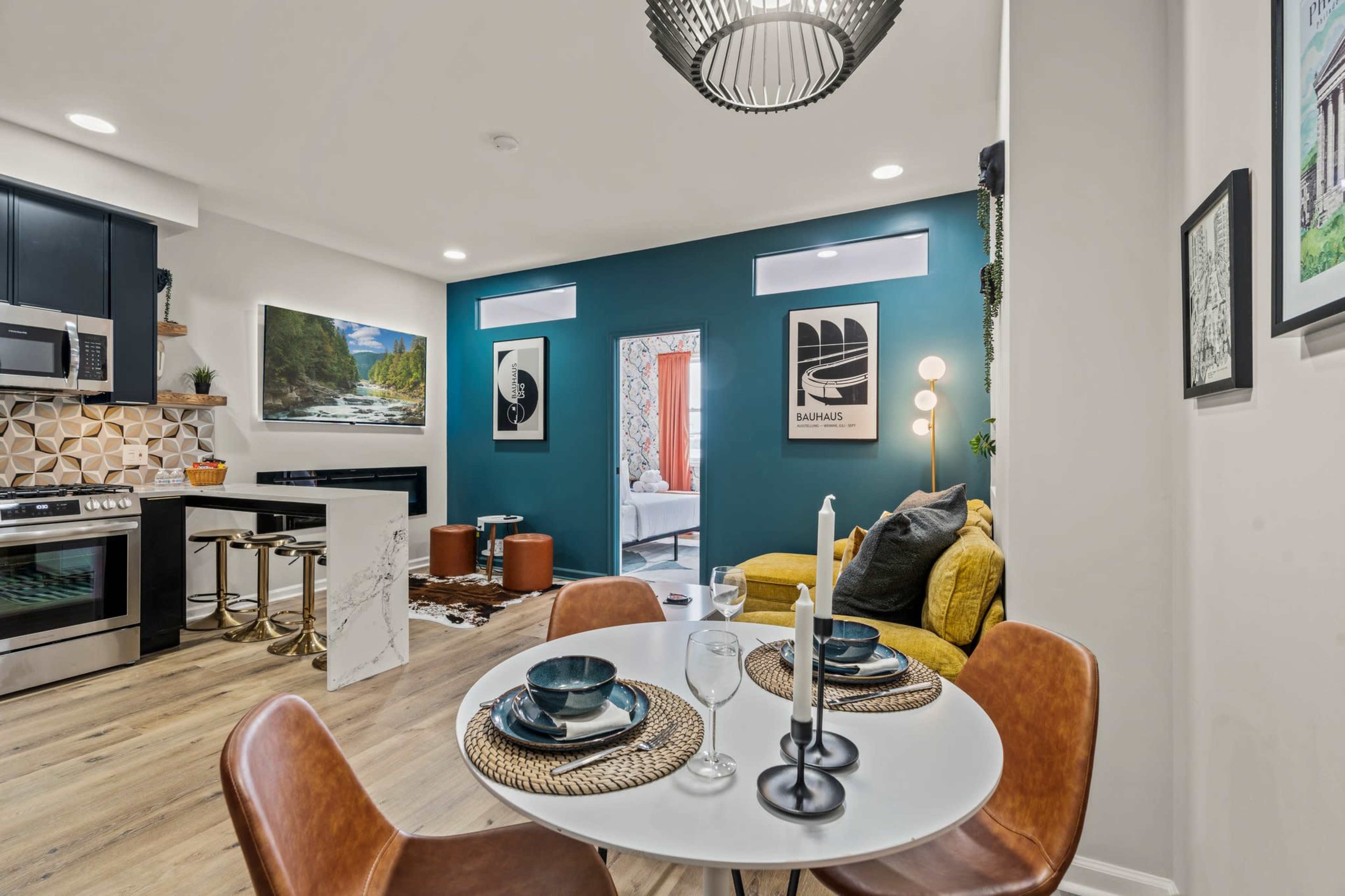 The image shows a modern kitchen and dining area featuring a circular table set for two, yellow seating, and a decorative wall in teal.
