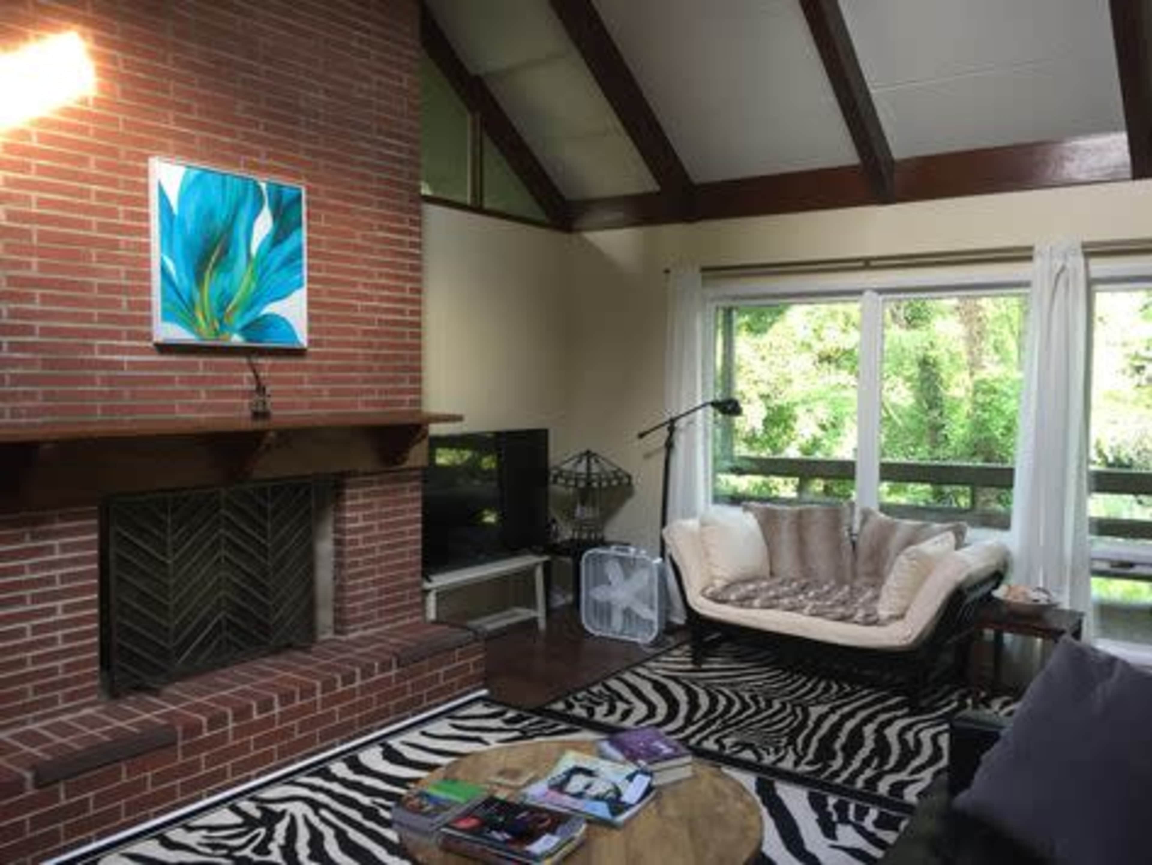 The image shows a living room with a brick fireplace, a large window overlooking greenery, a patterned rug, and a sectional sofa.