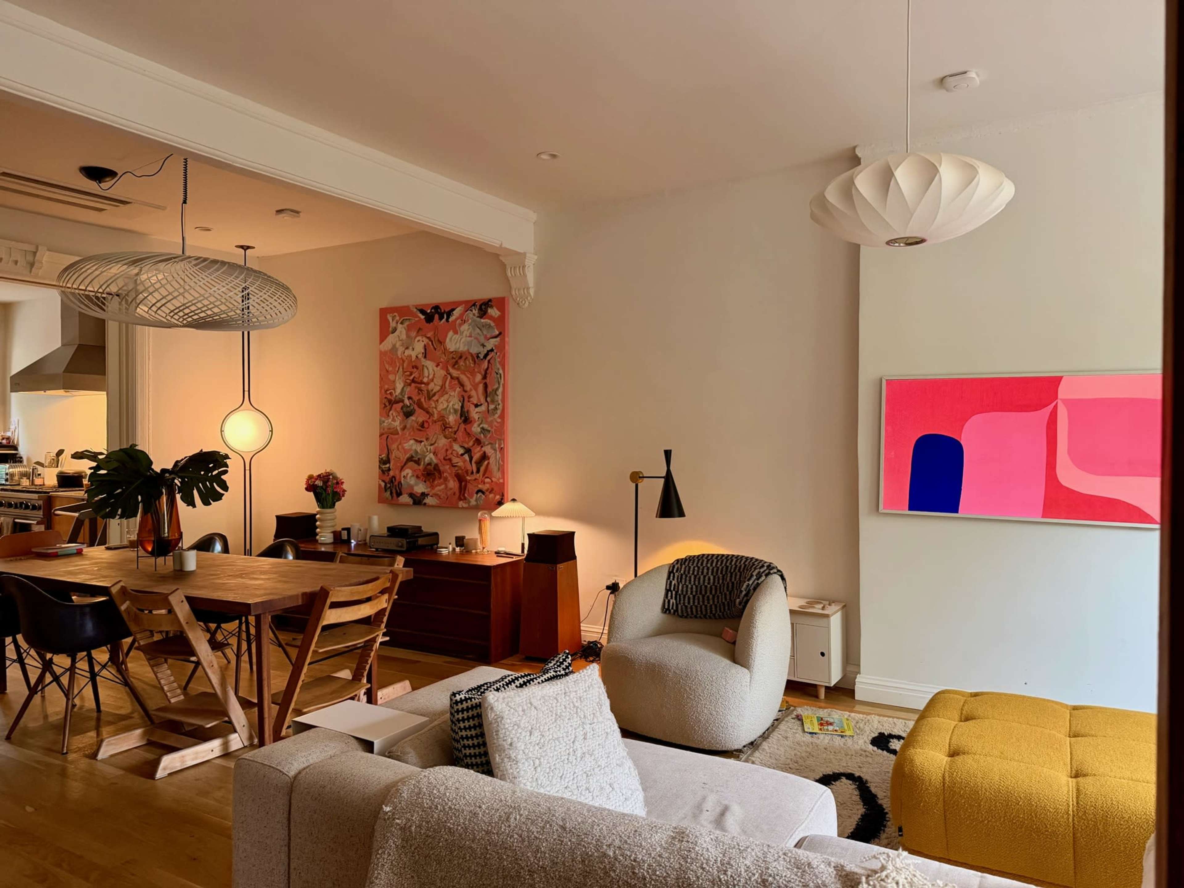 The image shows a modern living room with a cozy seating area, a dining table, decorative art on the walls, and a warm, inviting atmosphere.