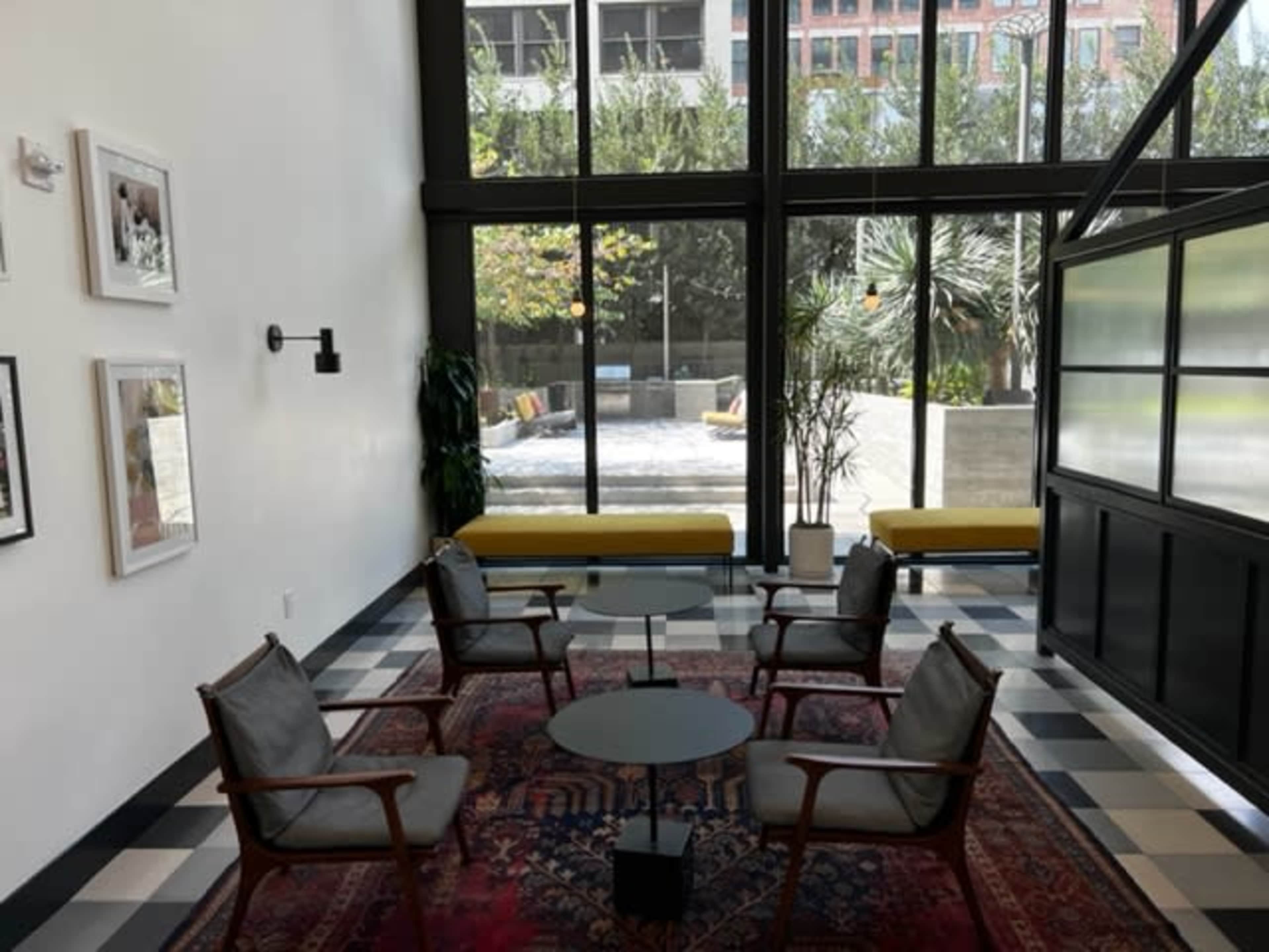 The image shows a modern indoor seating area with four wooden chairs and a glass table, surrounded by large windows that provide a view of an outdoor garden space.