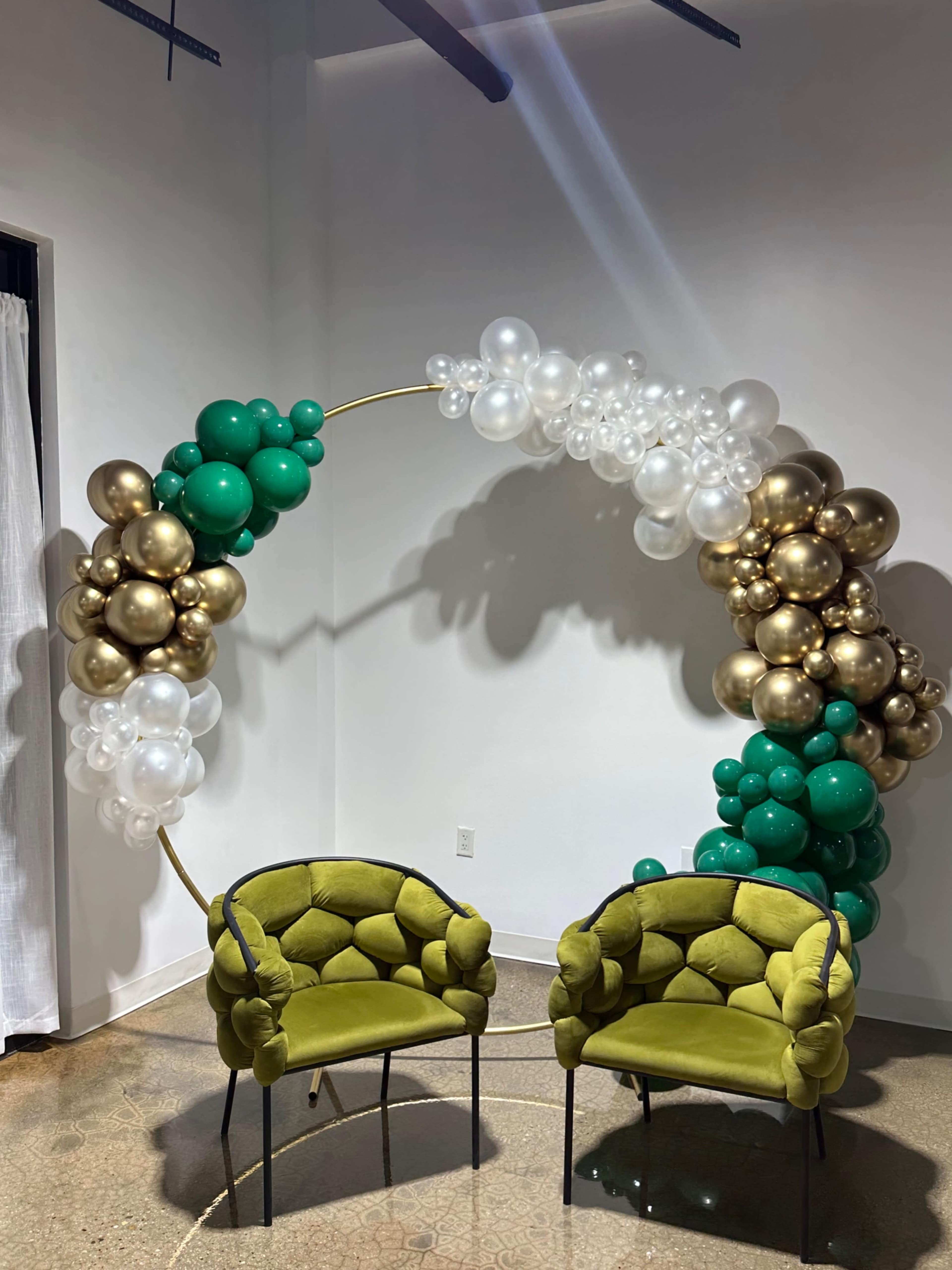 The image shows a decorative arch made of balloons in shades of green, gold, and white, positioned above two green upholstered chairs.