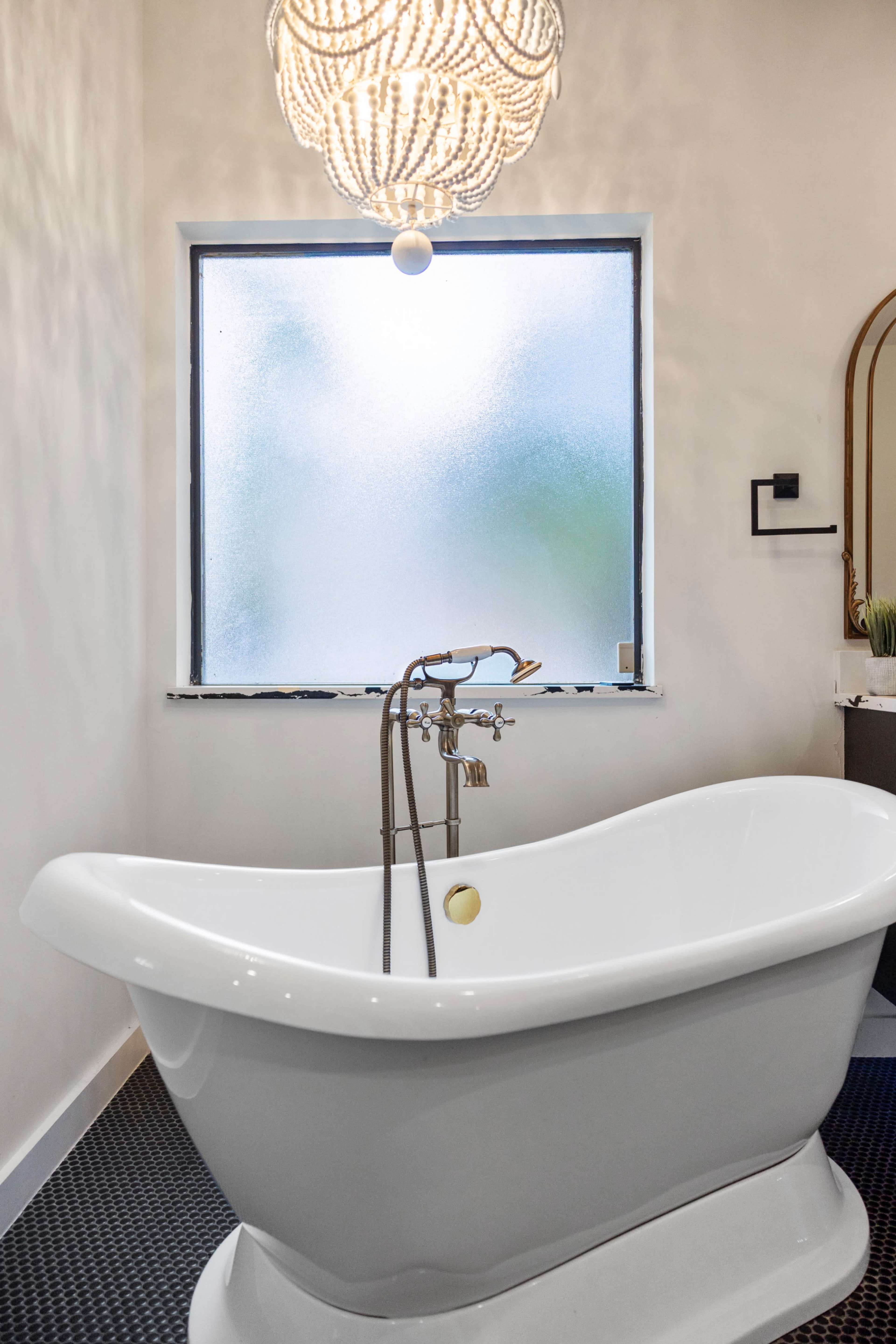 A freestanding white bathtub is positioned beneath a frosted window, with a decorative chandelier hanging above in a modern bathroom.