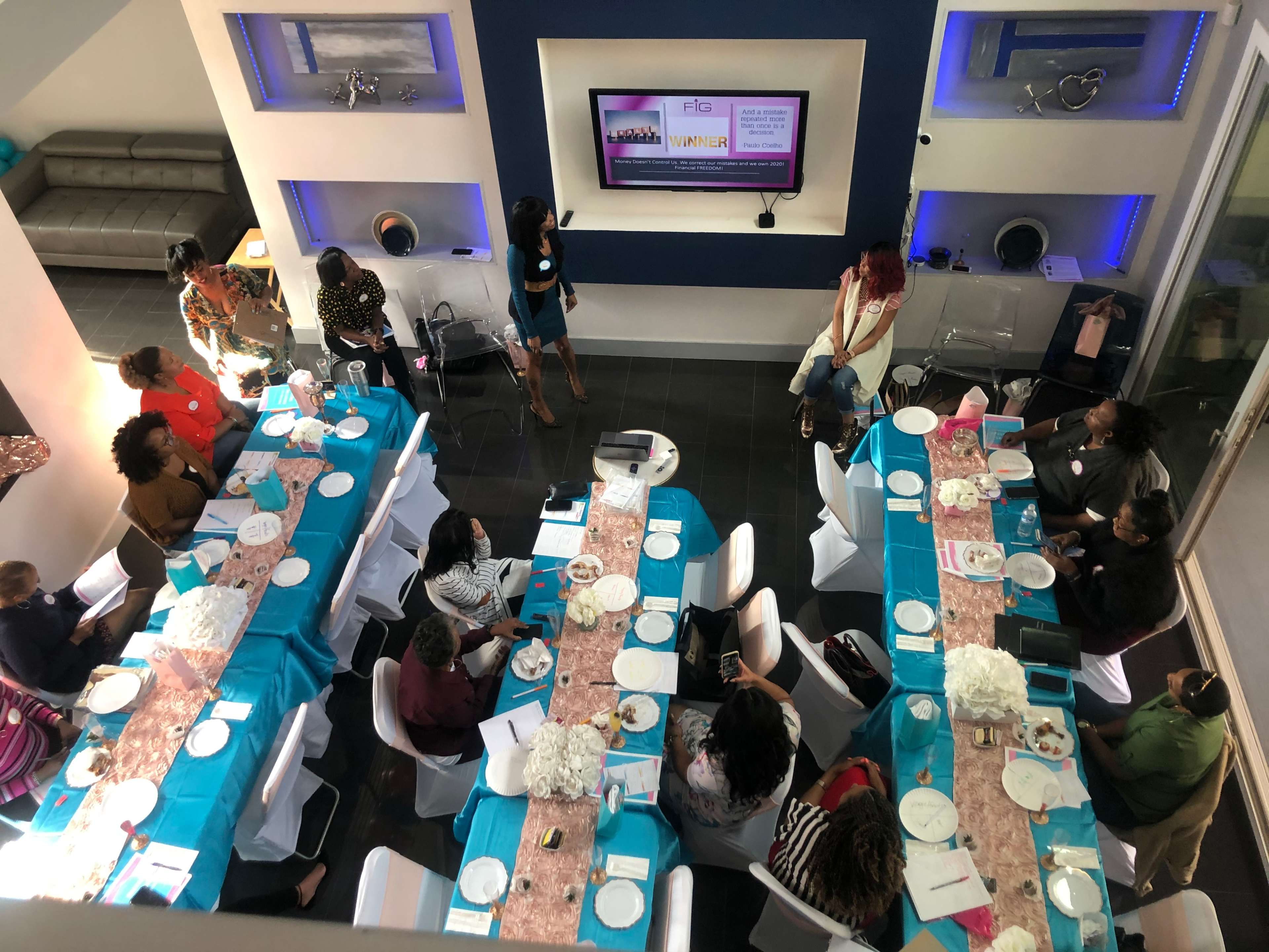 A group of people is seated at a long table covered with turquoise tablecloths in a modern room, with a presentation screen displaying information at one end.