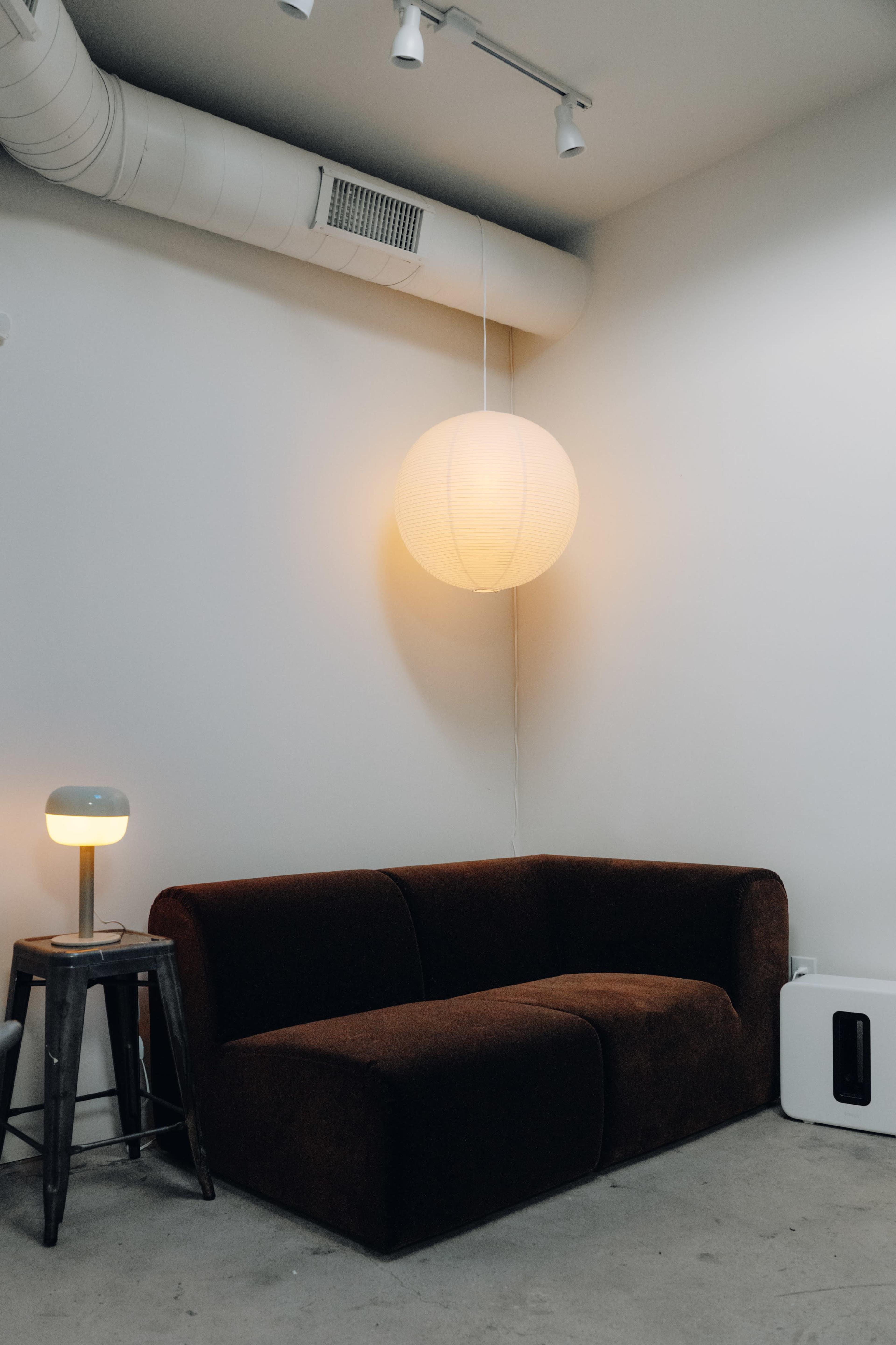 The image shows a simple living room setup featuring a brown sofa, a small lamp on a wooden stool, and a round hanging light fixture against a white wall.