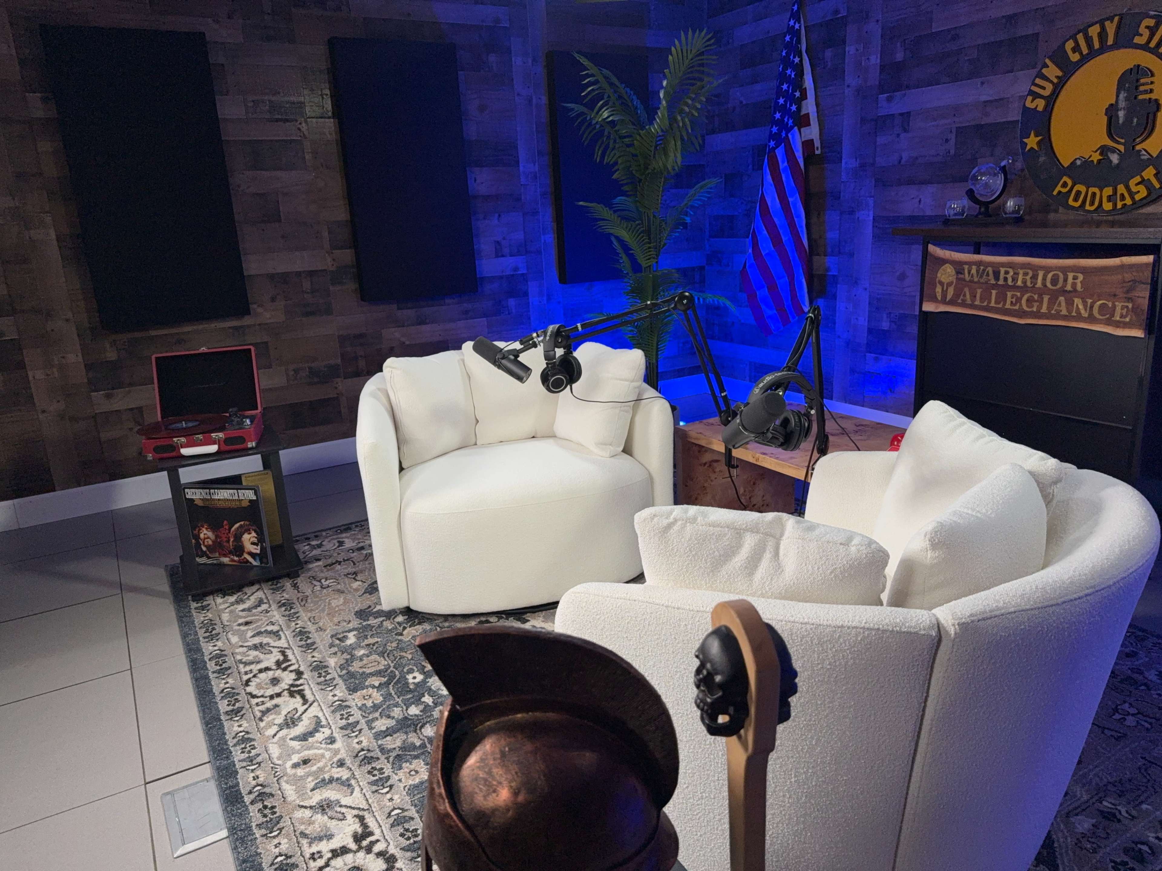 The image depicts a cozy podcast recording studio featuring two white armchairs, a wooden table, and a backdrop with an American flag and a sign that reads "Warrior Vigilance."