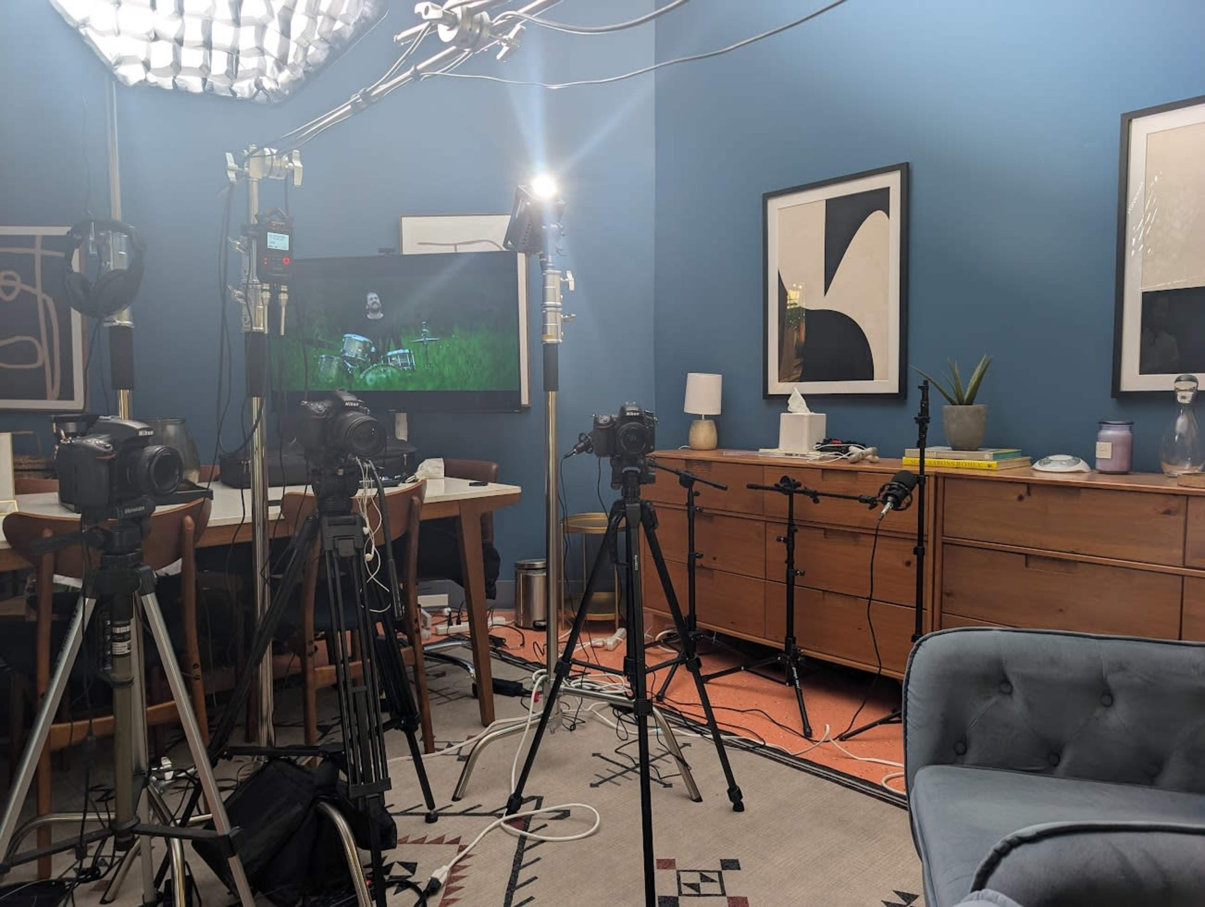 The image shows a room equipped for filming, featuring multiple cameras on tripods, lighting equipment, and a large screen displaying an image, with a mix of furniture and decor in the background.