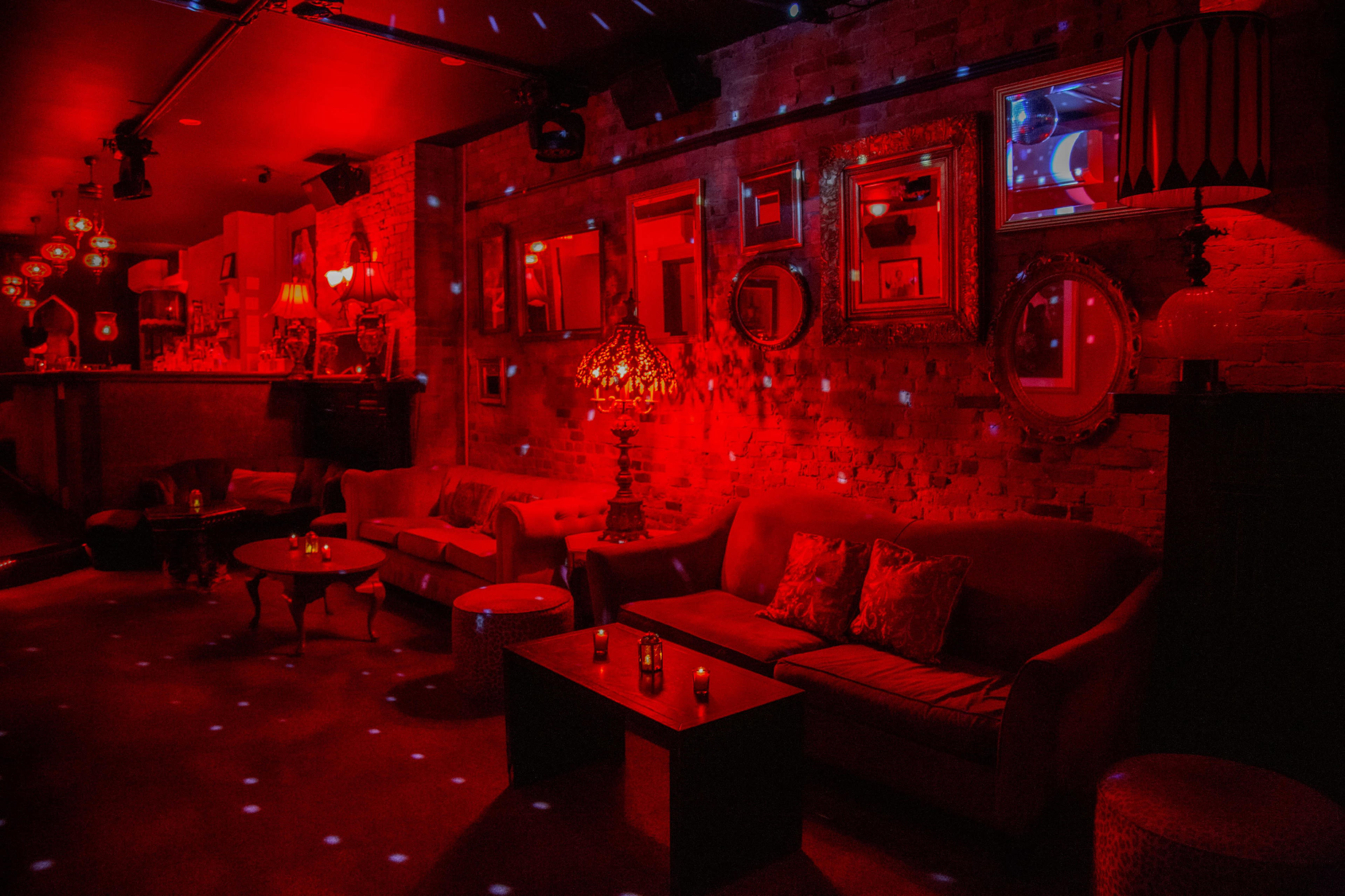 The image shows a dimly lit lounge area with red lighting, featuring sofas, a coffee table, and multiple mirrors on the walls.