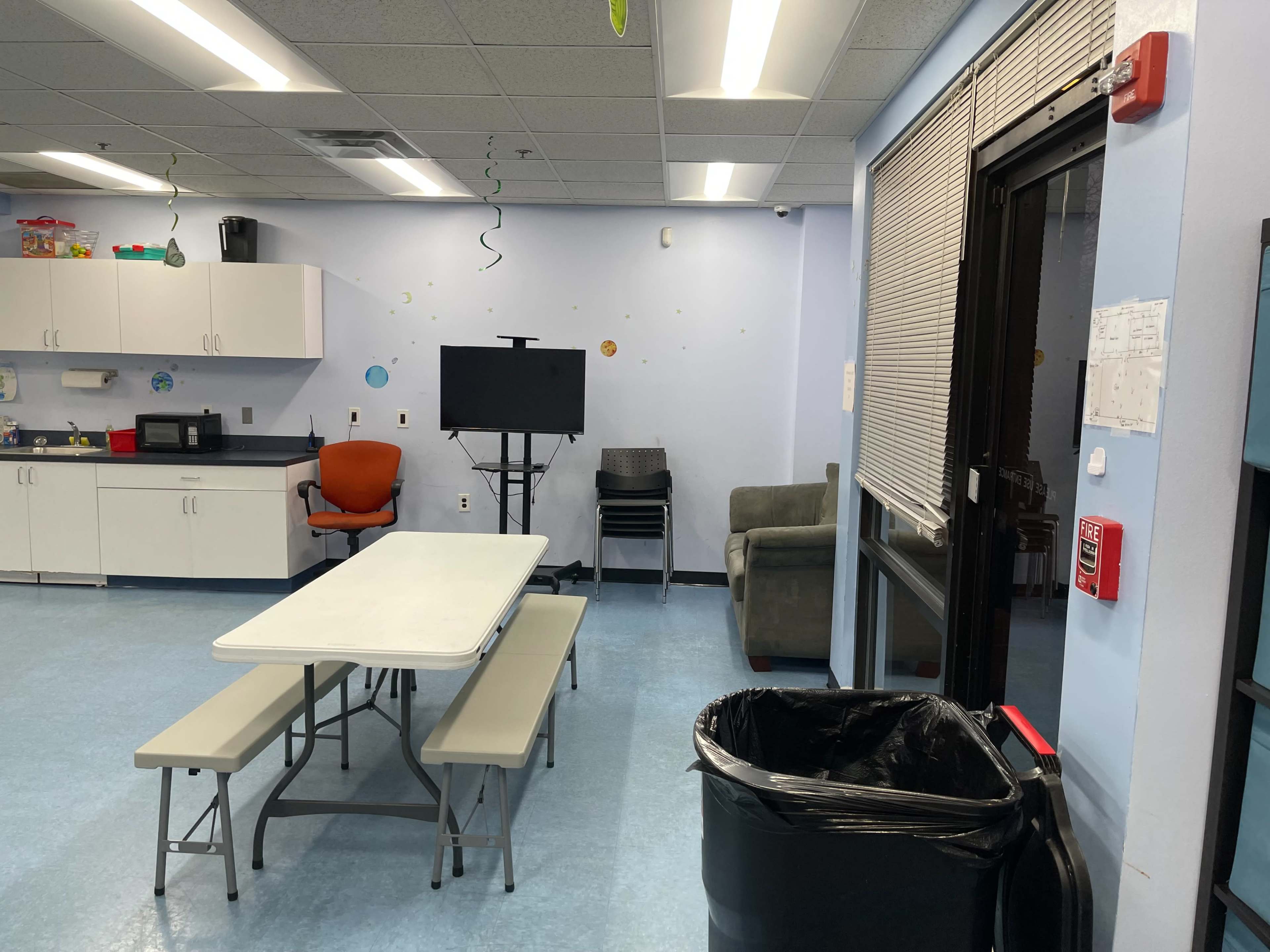 A room with a kitchen area featuring a table with benches, a microwave, a television on a stand, a stack of chairs, and a trash bin.