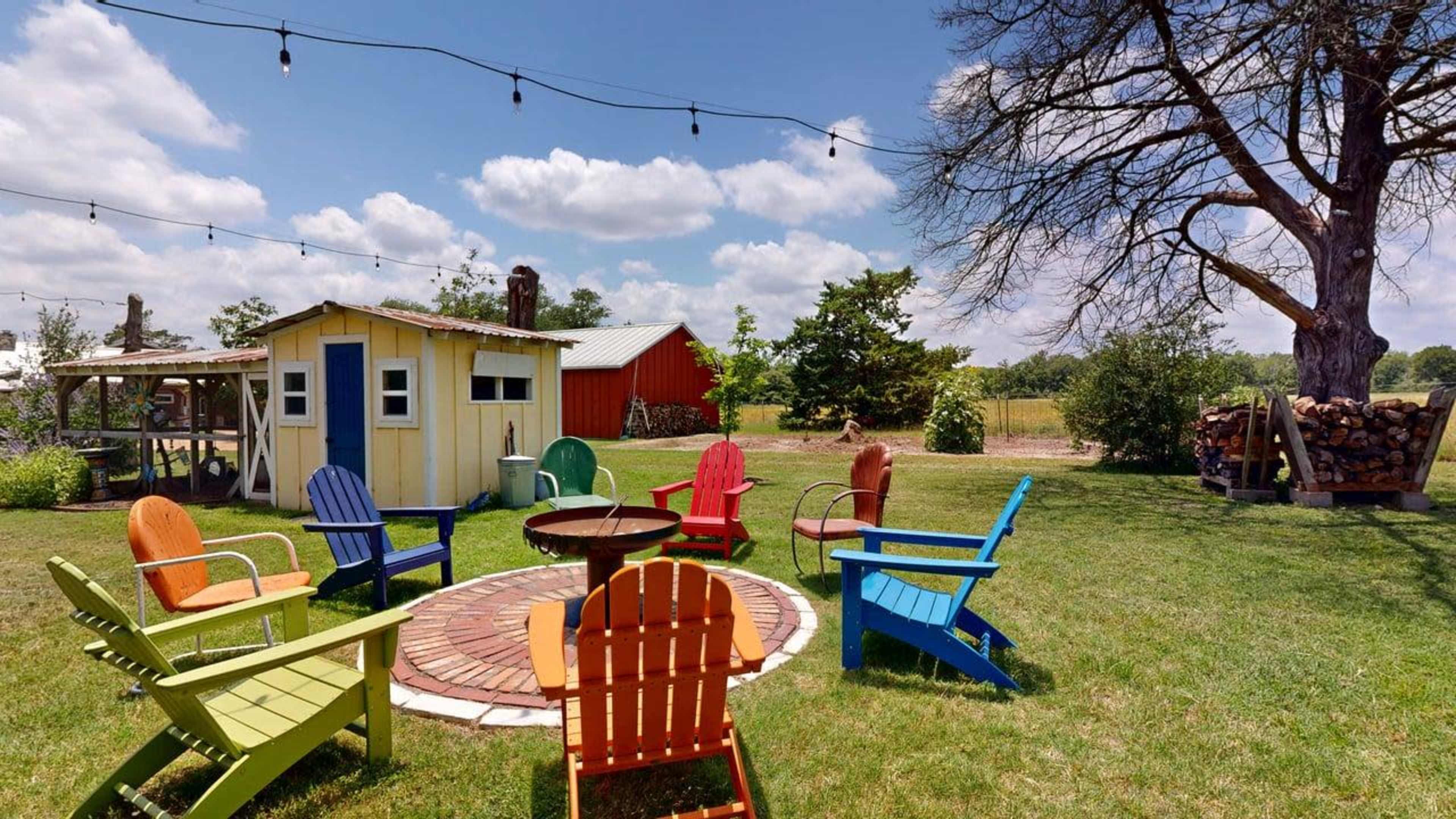A circular arrangement of brightly colored Adirondack chairs surrounds a fire pit in a grassy backyard, with a small shed and a red barn in the background under a partly cloudy sky.