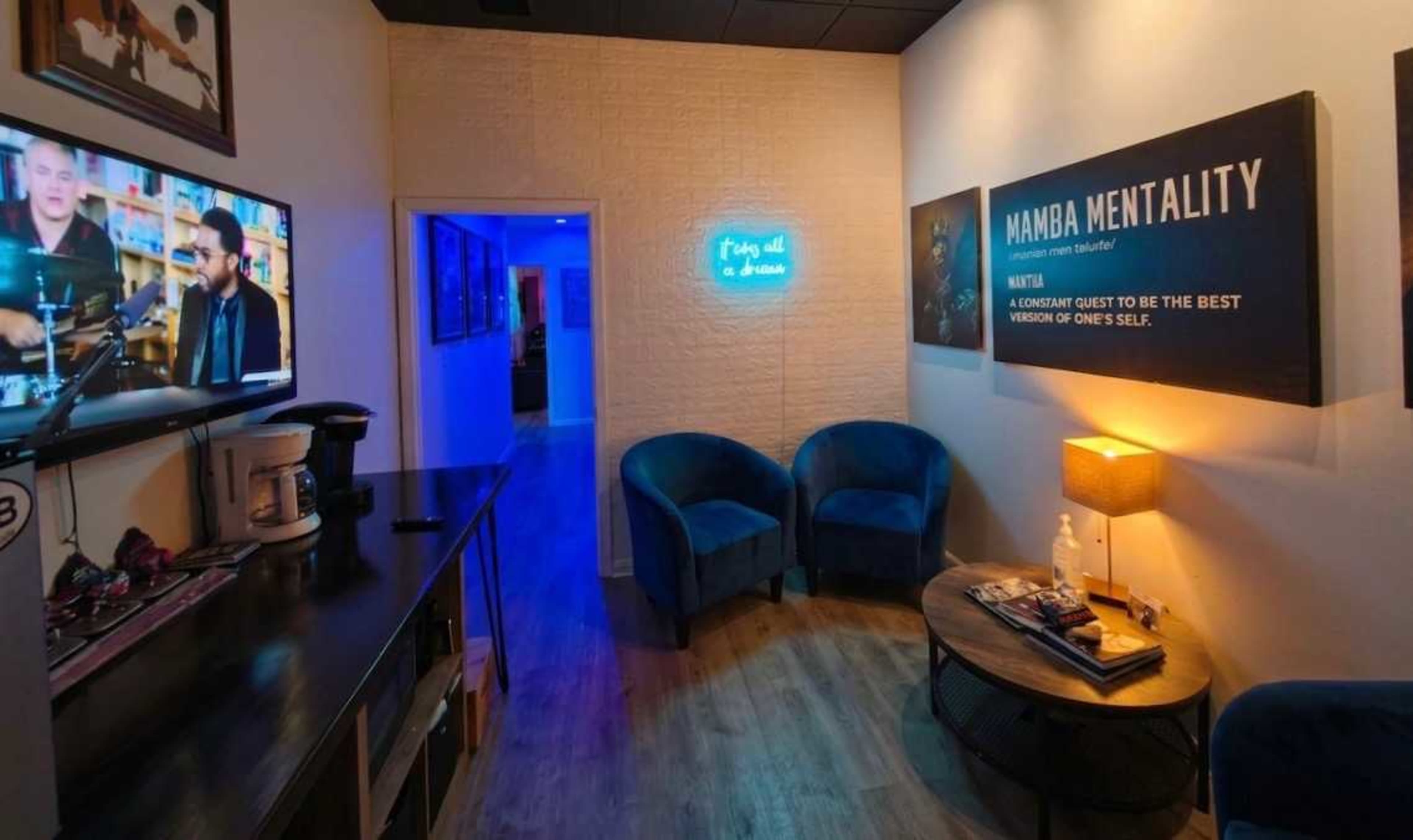 The image shows a cozy lounge area featuring two blue chairs, a small round table, a television displaying a video, and motivational wall art illuminated by soft blue lighting.
