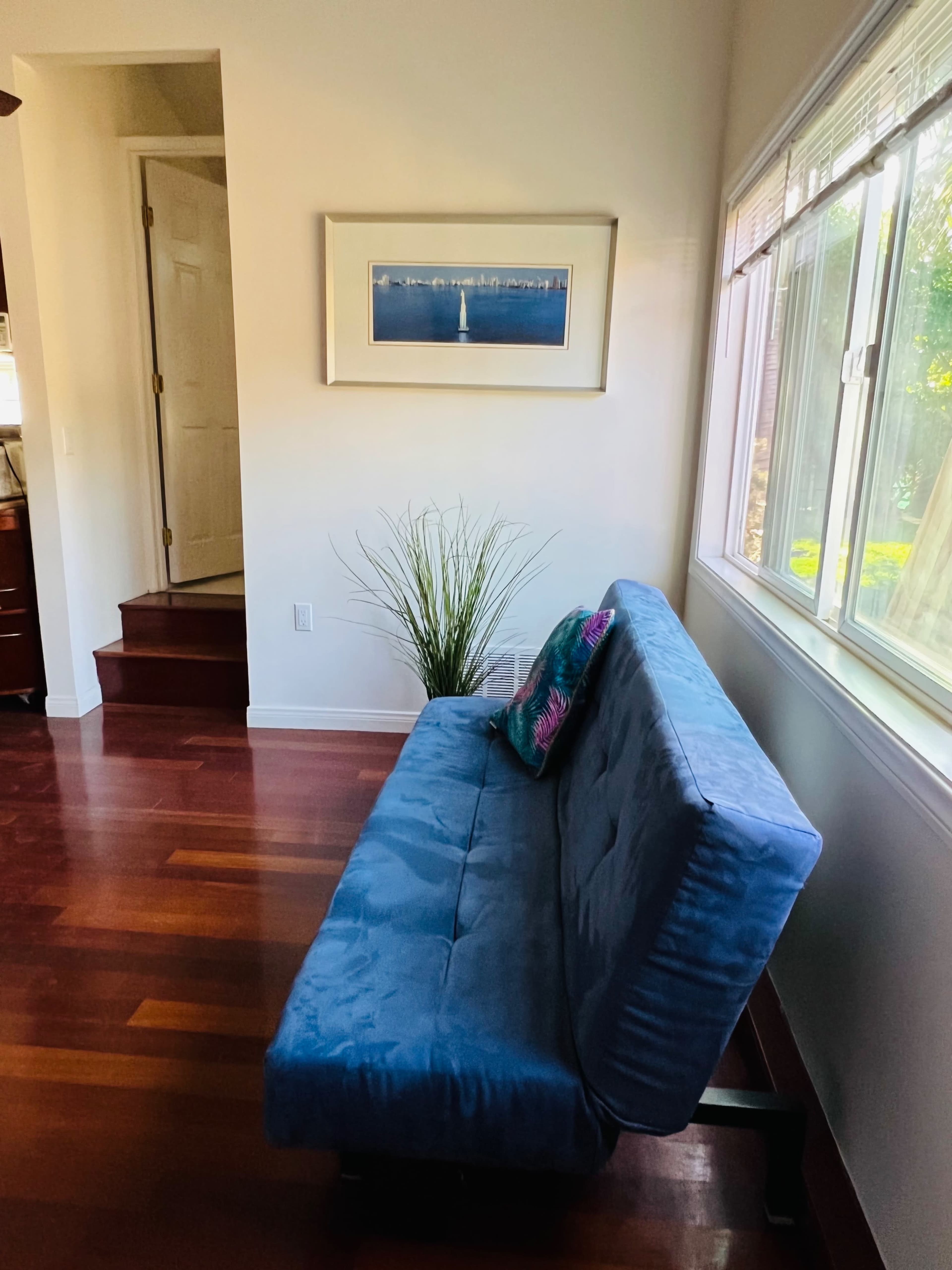 A blue sofa against a wall next to a window with a framed picture above it and a potted plant nearby.