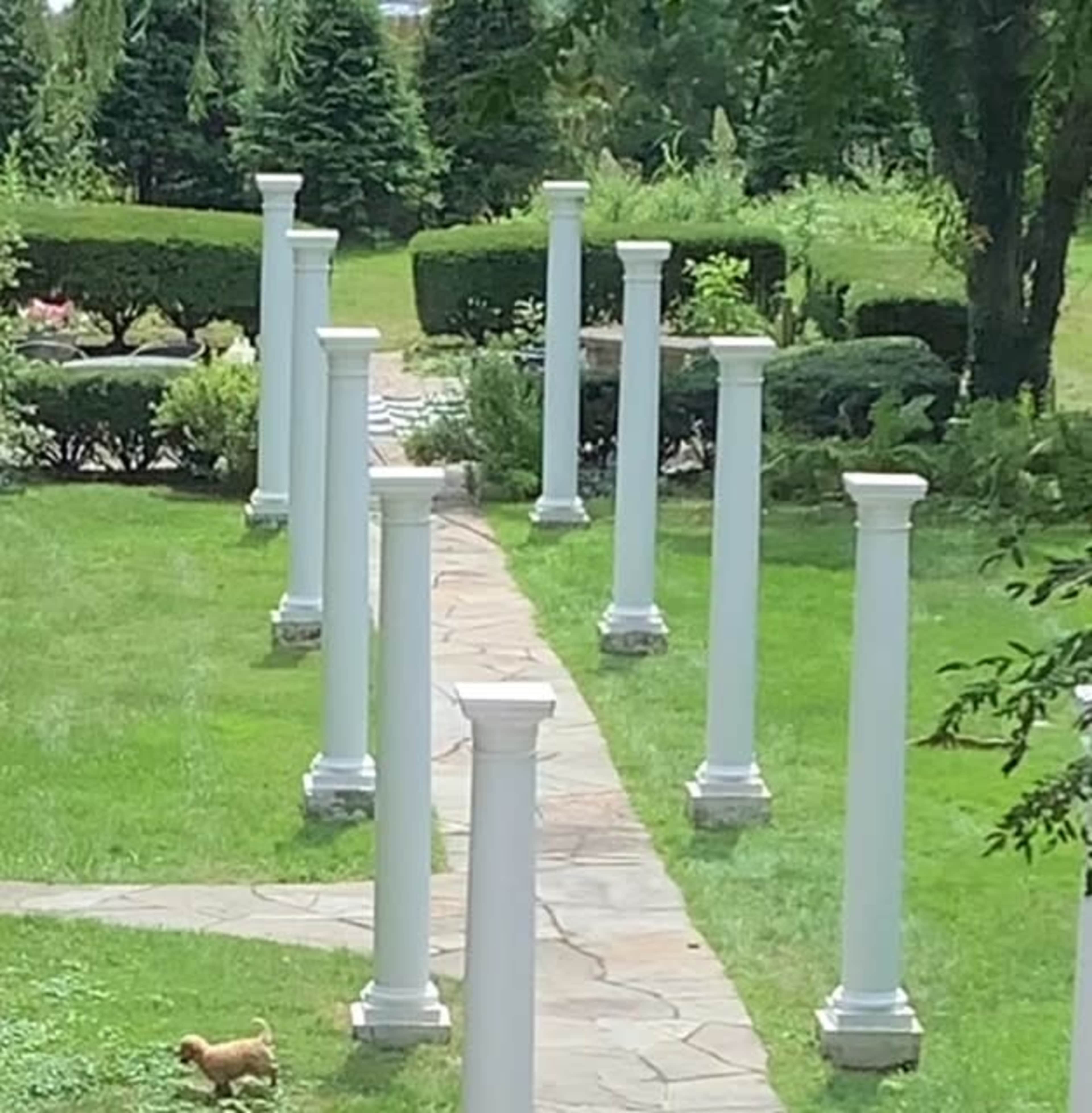 A paved pathway lined with white columns runs through a grassy area with neatly trimmed bushes and trees.