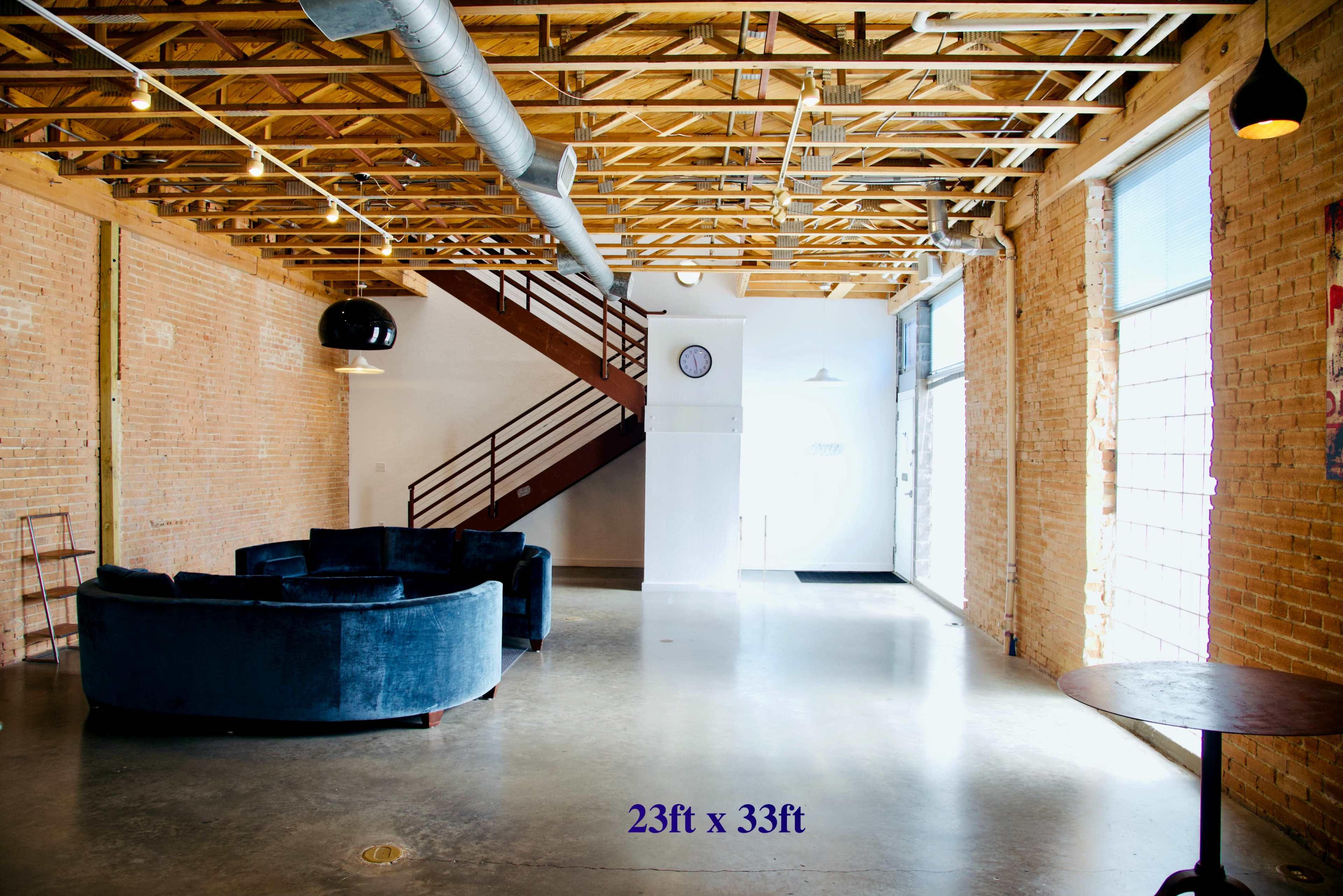 A spacious interior with a large, circular blue sofa, a wooden staircase in the background, and large windows providing natural light.