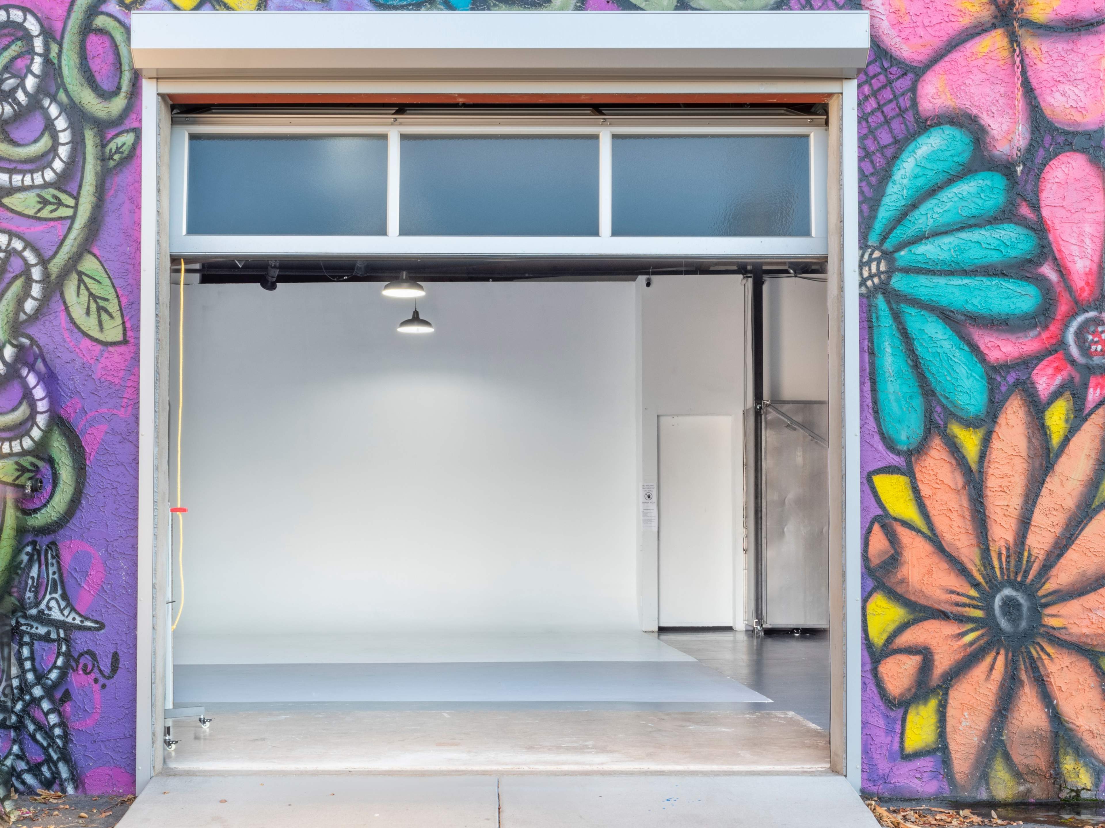 A large open garage door reveals a minimal interior space with plain walls and a polished floor, framed by vibrant graffiti on the outside.