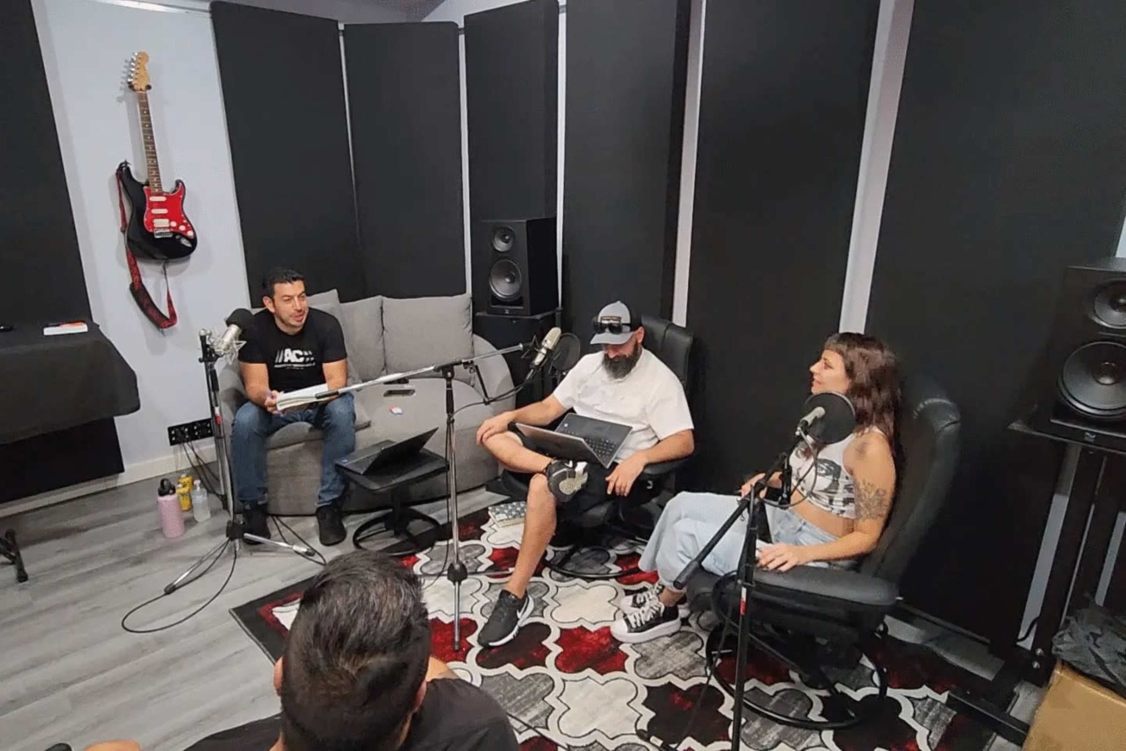 A group of three people is seated in a recording studio, discussing topics while microphones are positioned in front of them.