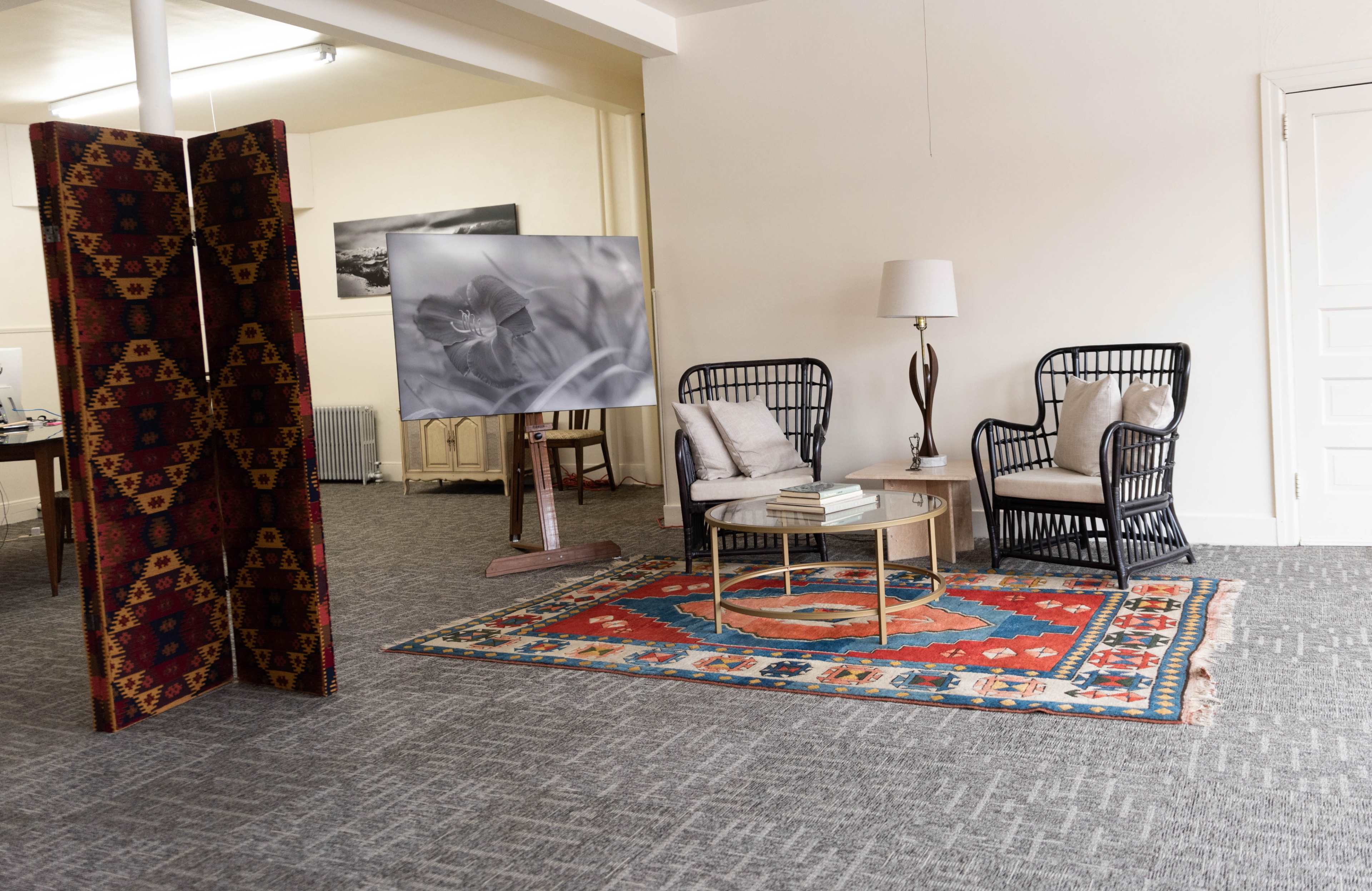 A cozy seating area with two wicker chairs around a round coffee table, set on a patterned rug, with a large framed photo and a decorative folding screen nearby.
