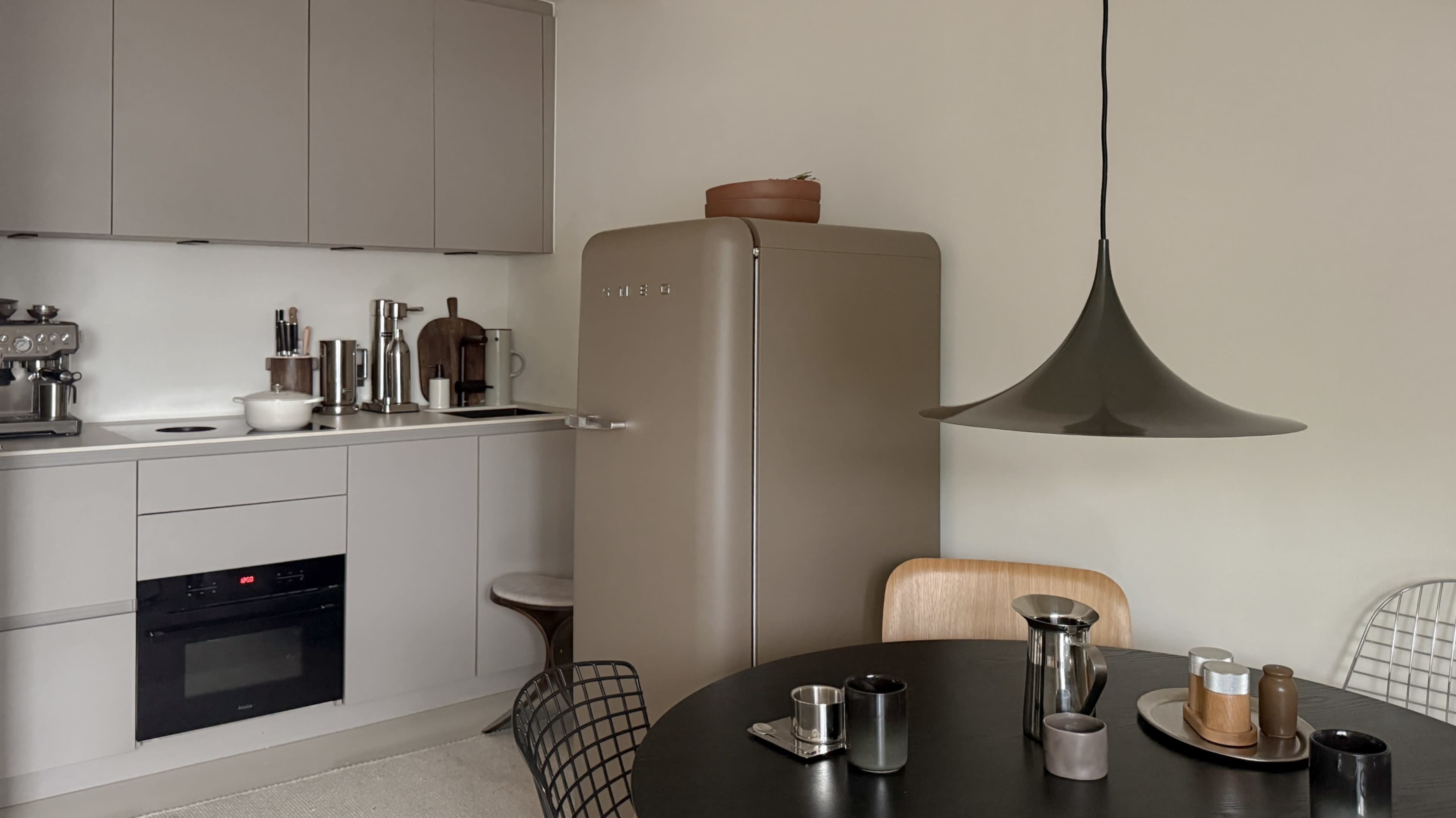 The image shows a modern kitchen with gray cabinets, a beige refrigerator, a round black dining table, and a pendant light overhead.