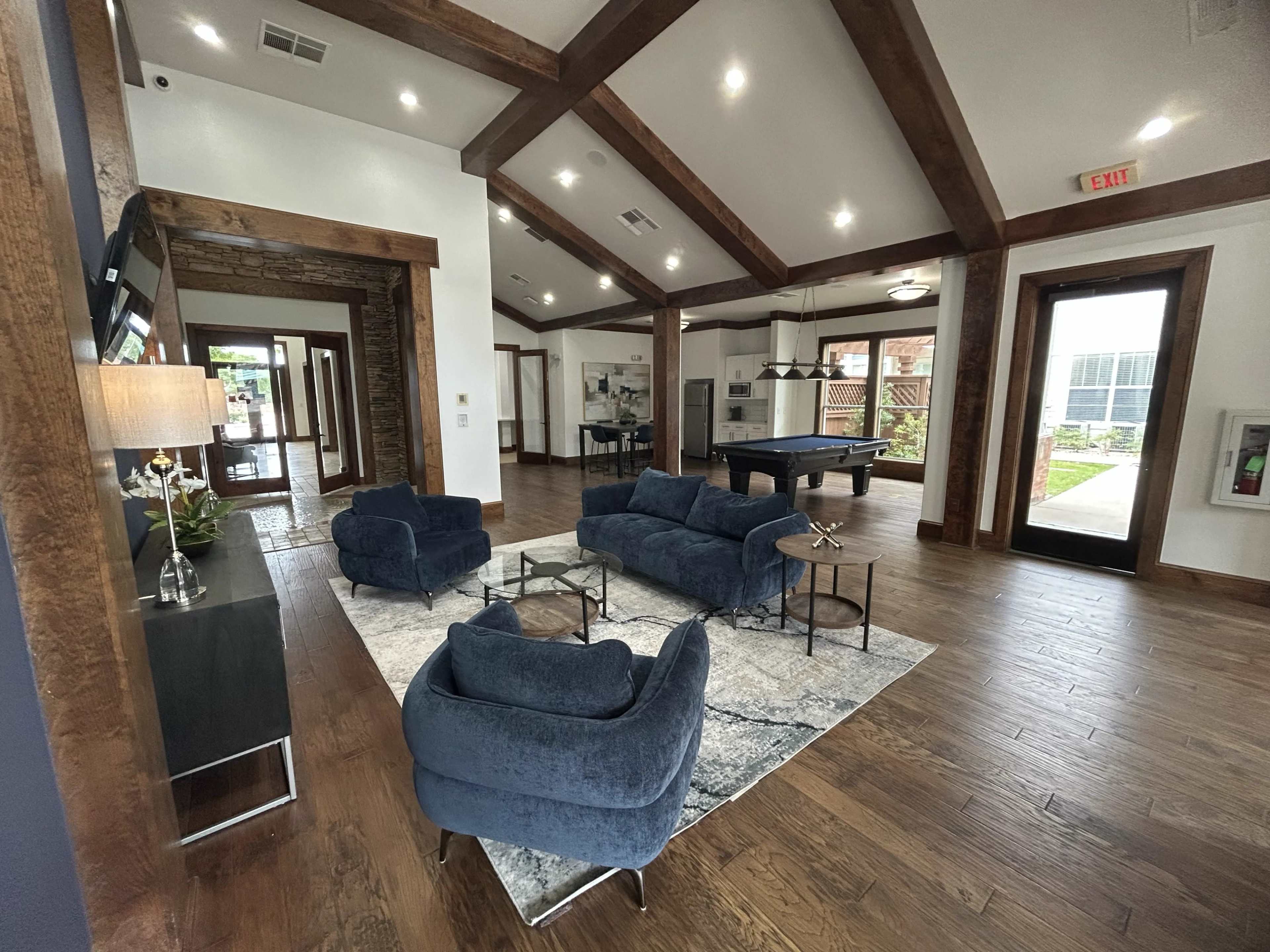 The image shows a modern, spacious common area with dark wooden beams, a pool table, and various seating arrangements on a large area rug.