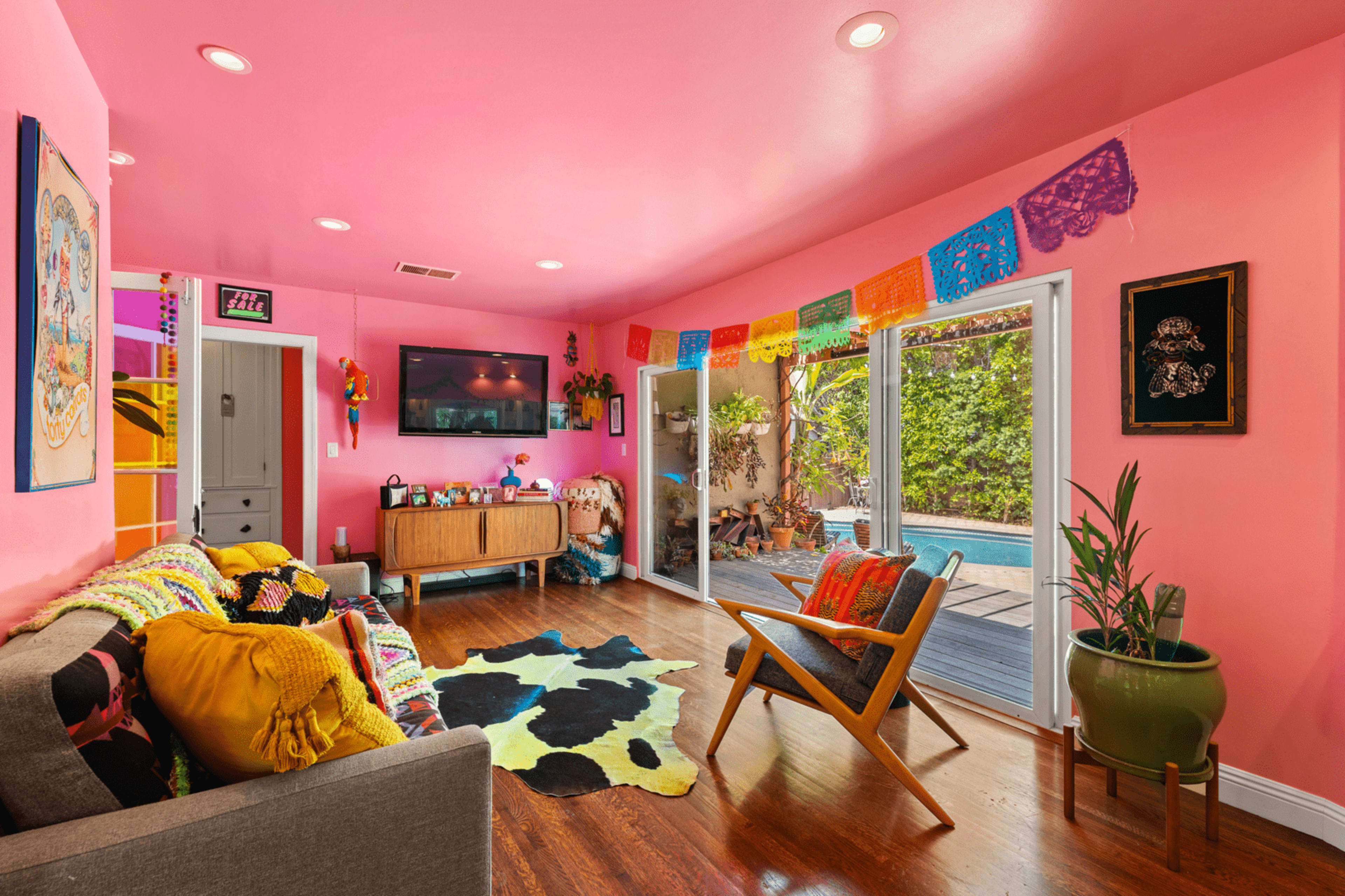 A bright pink living room features a gray couch, colorful wall decorations, and large windows overlooking a pool area.
