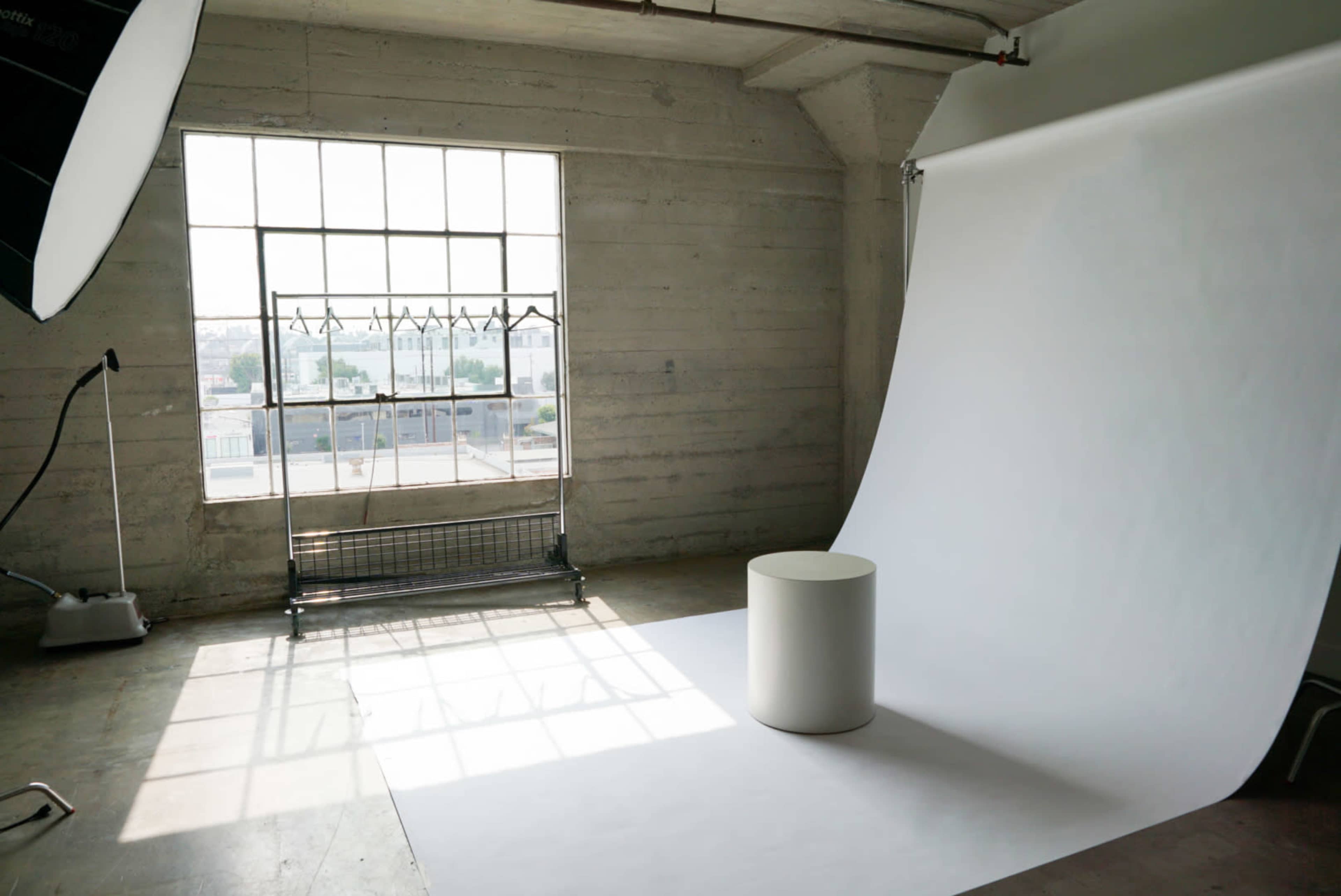 The image shows a photography studio featuring a large window, a white backdrop, and a round pedestal in front of the backdrop.