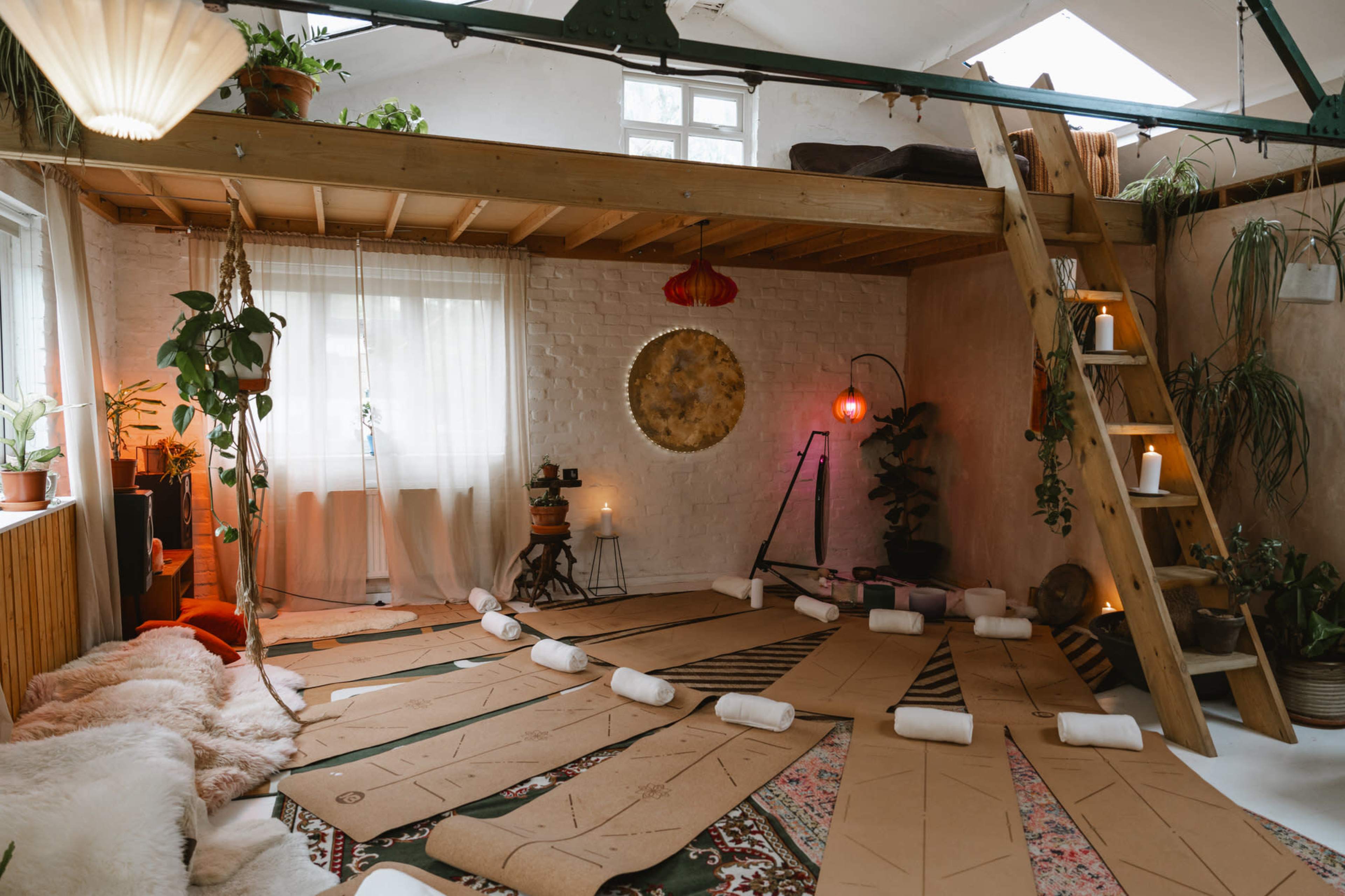 The room features a lofted area with wooden beams, a circular arrangement of brown paper, and various plants adorning the space.