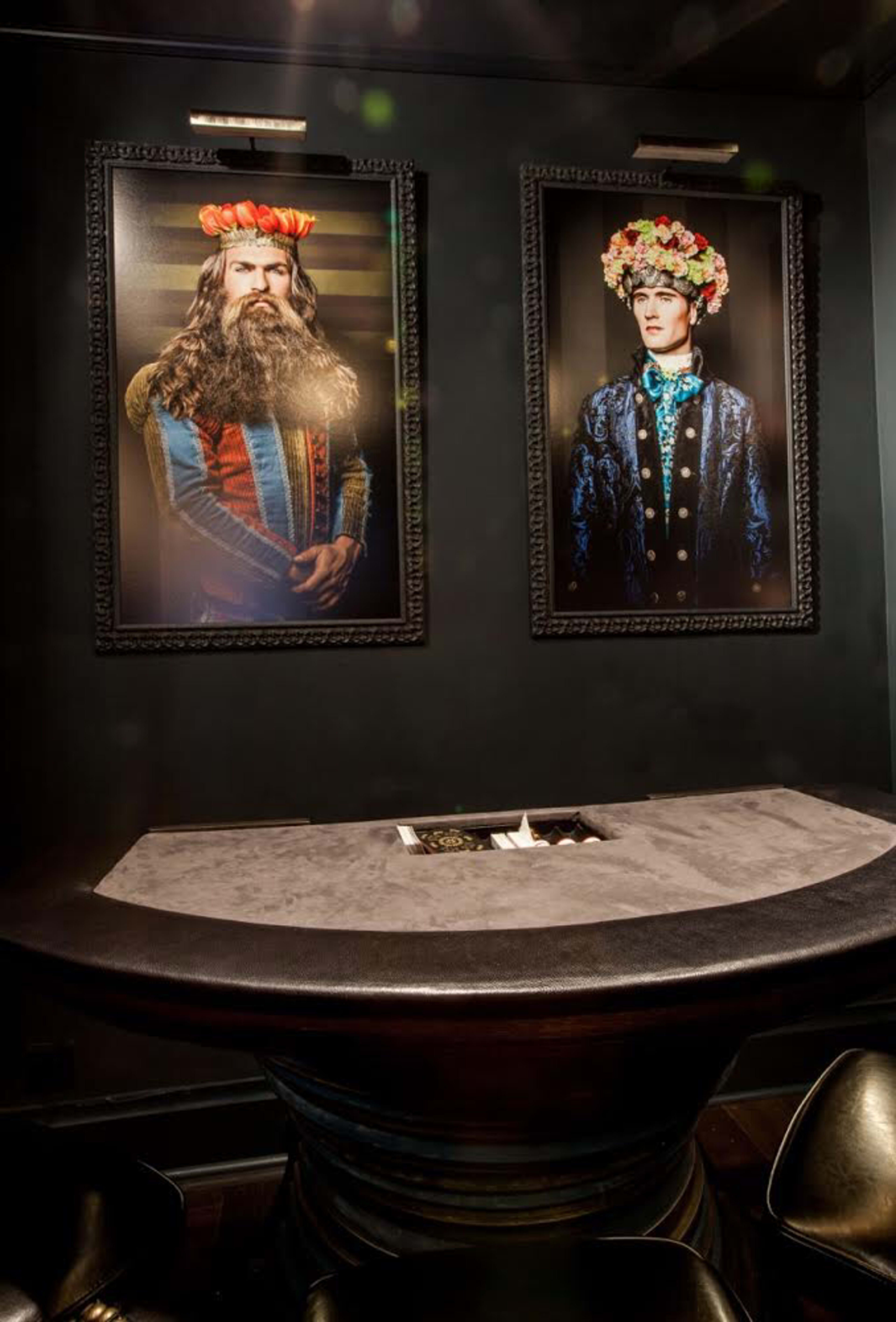 Two large framed portraits of individuals with floral headpieces displayed on a dark wall above a round table with a fire pit in the center.