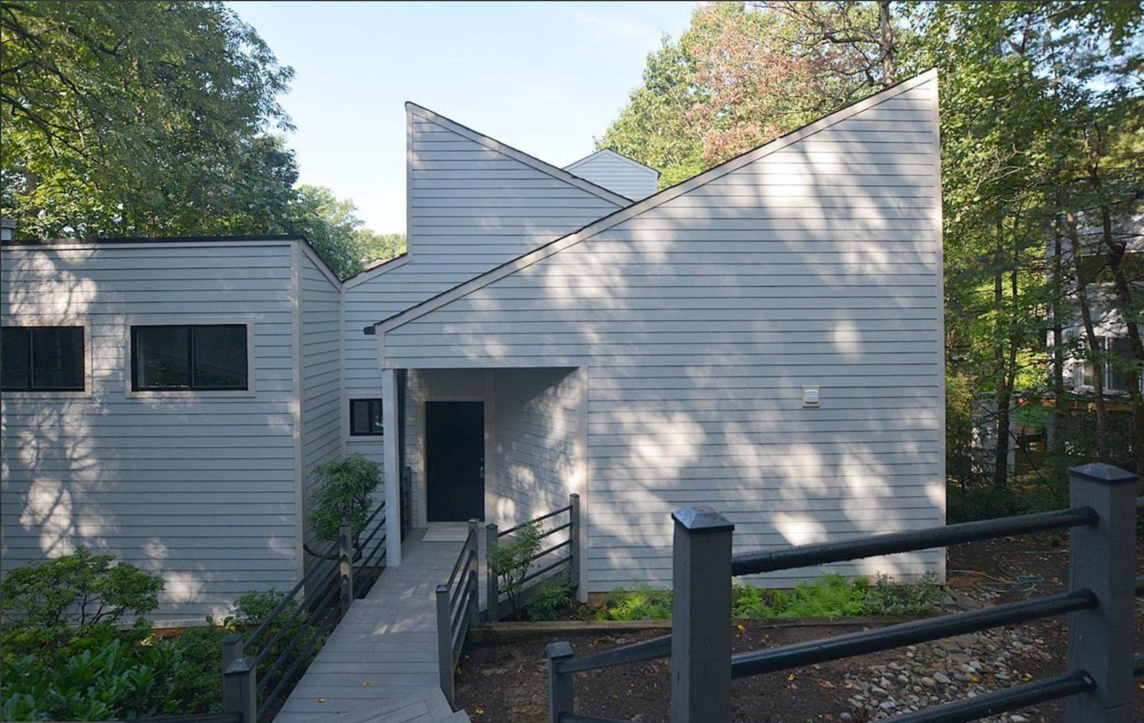 The image shows a modern, angular house with gray siding and a pathway leading to a black front door surrounded by trees.